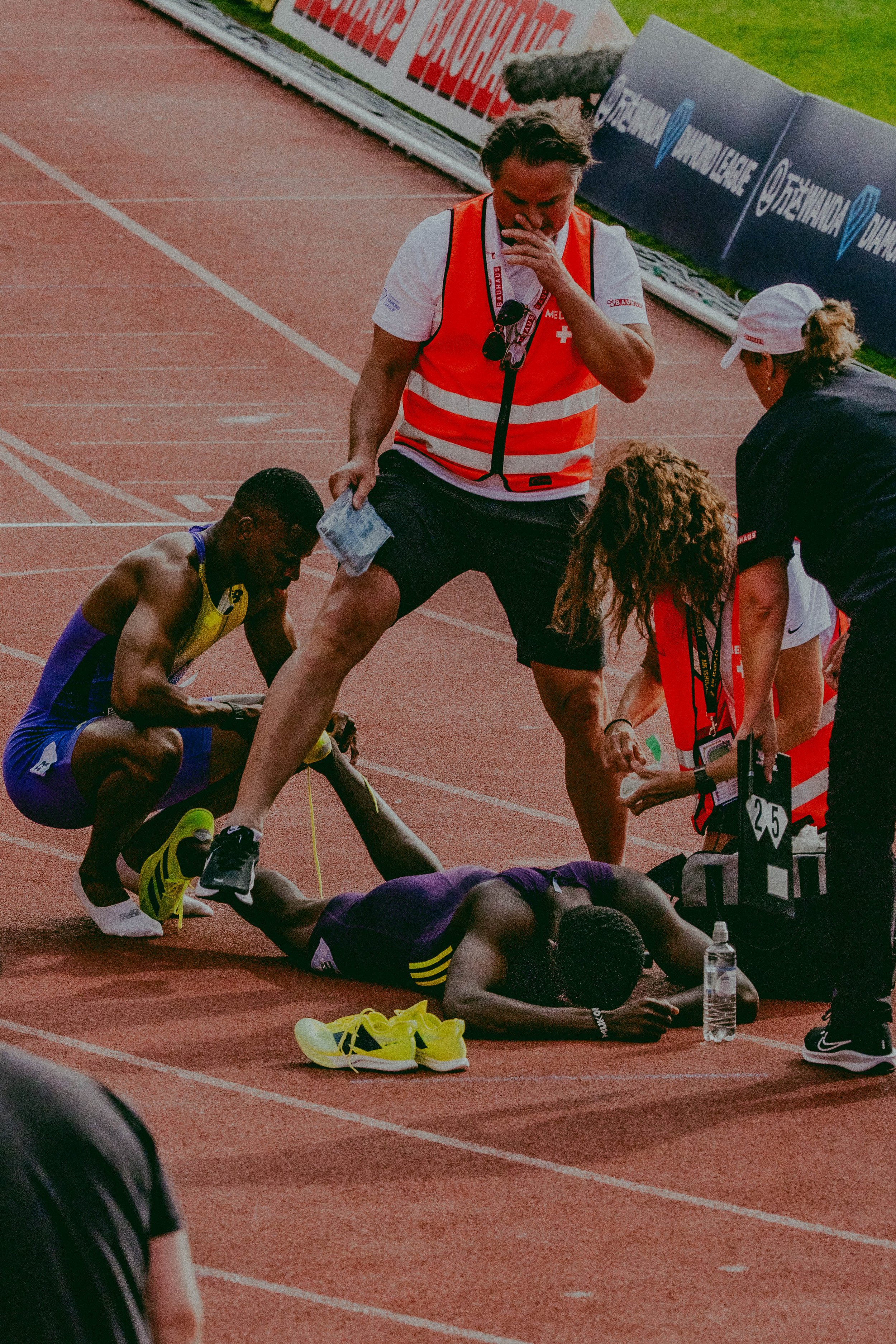 A marathon runner appears to have collapsed on the track, with medical personnel attending to him. A woman in a white cap and dark jacket is crouched nearby, possibly providing assistance. The background shows a running track and advertising banners.