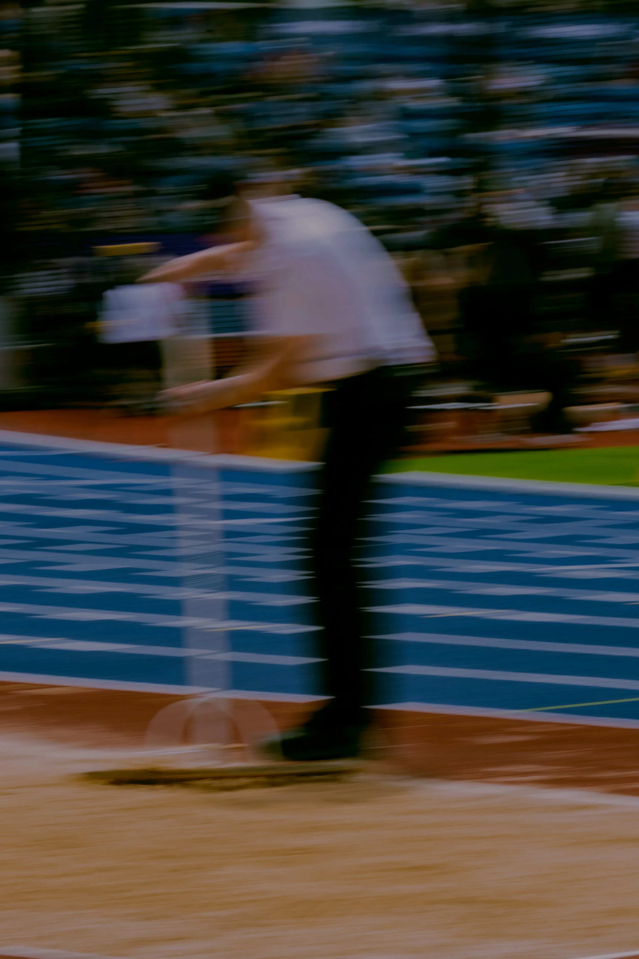 Blurred photo of a person playing tennis on an outdoor court, with a crowd watching in the background.