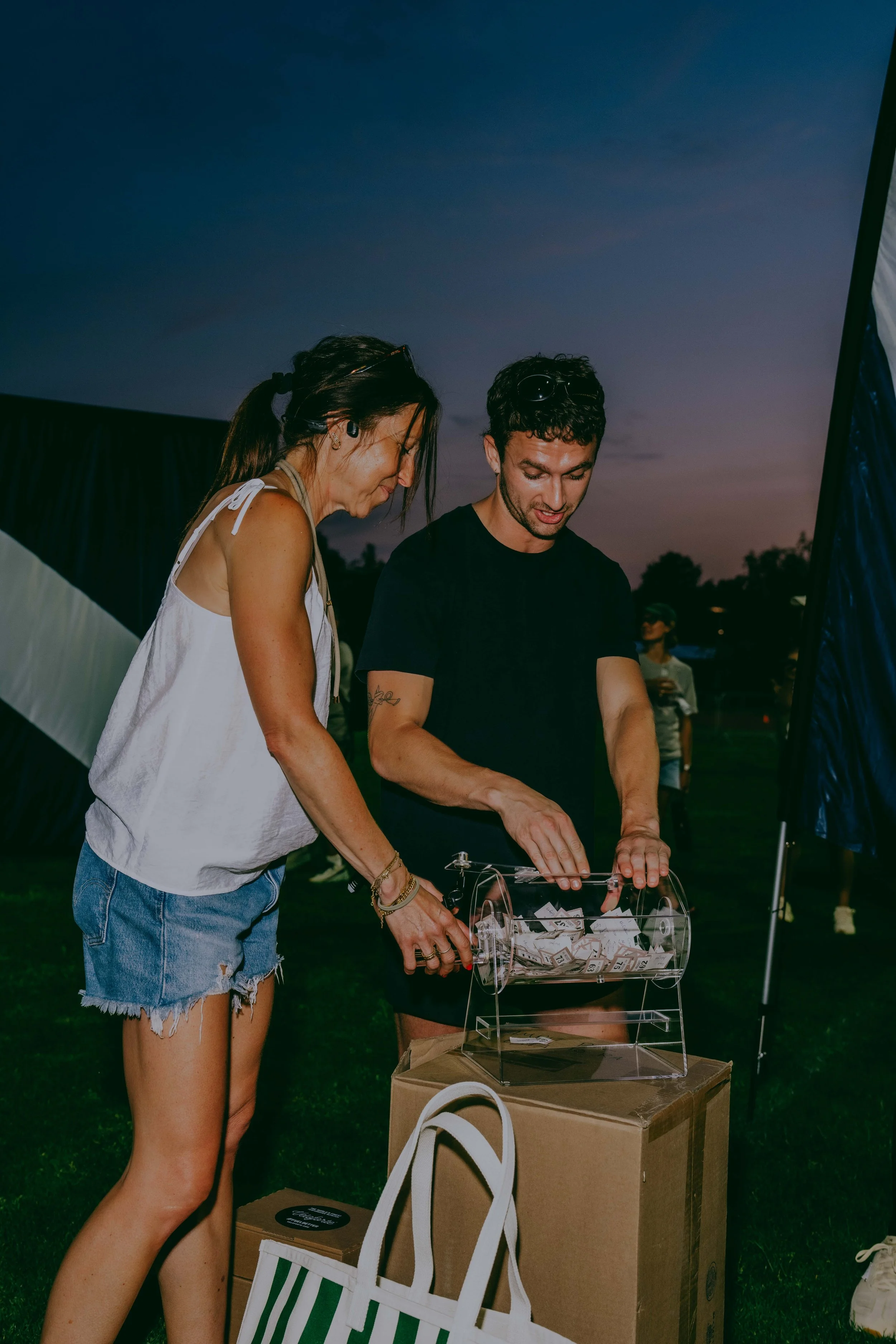 Two women at an outdoor event are drawing tickets from a transparent box filled with paper slips. One woman is wearing a white sleeveless top and denim shorts, while the other is in a black t-shirt. The sky is darkening as evening approaches. There a