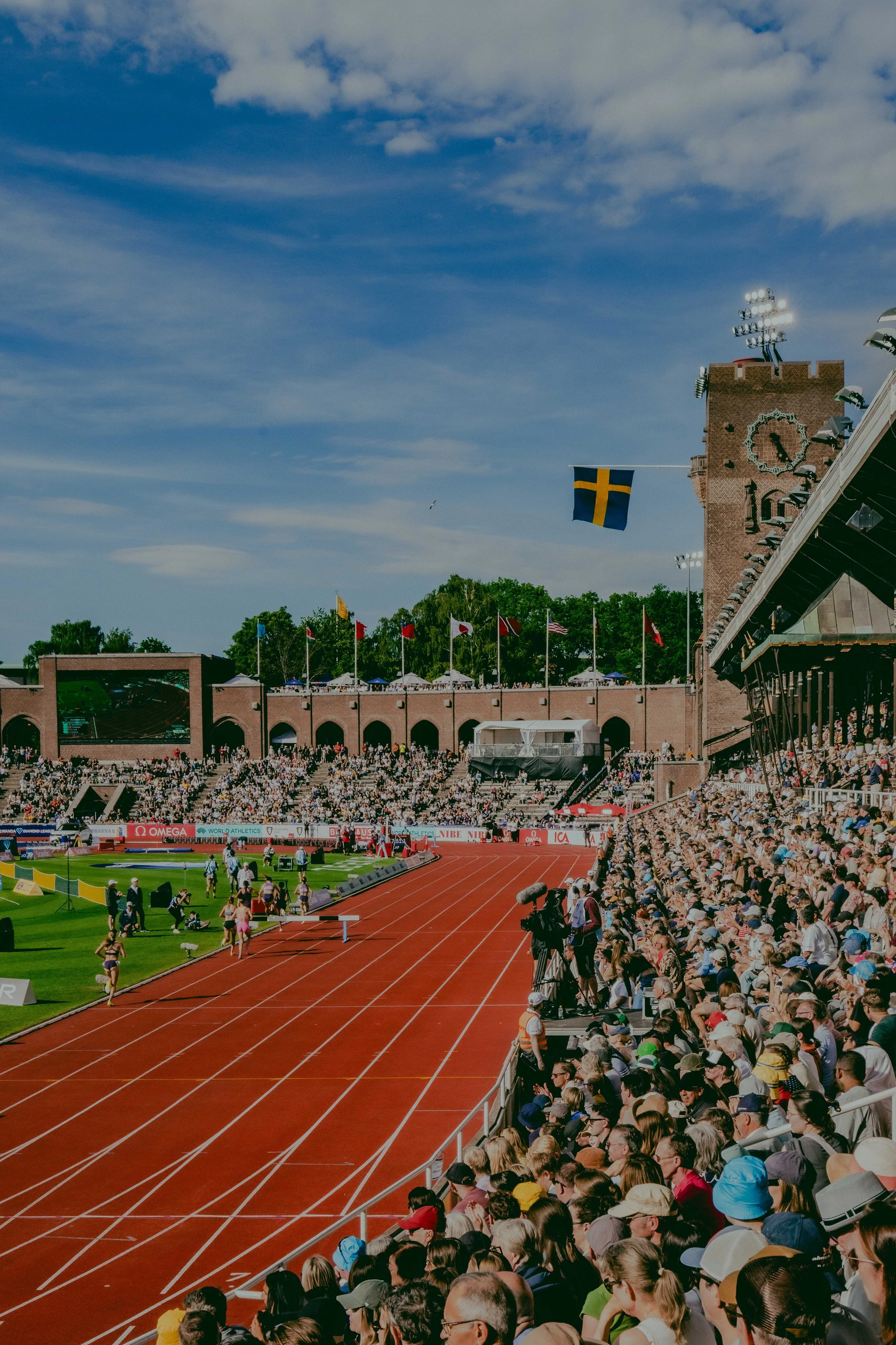 A crowded outdoor track and field stadium during a sporting event, with many spectators seated in the bleachers and a large Swedish flag flying near a castle-like structure, under a partly cloudy sky.