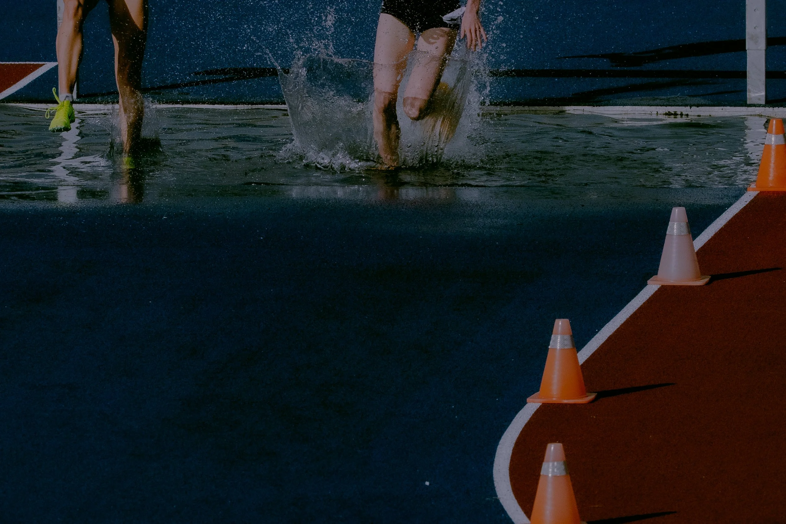 Close-up of two athletes running on a wet track, with orange cones marking the lane, and water splashing as they run.