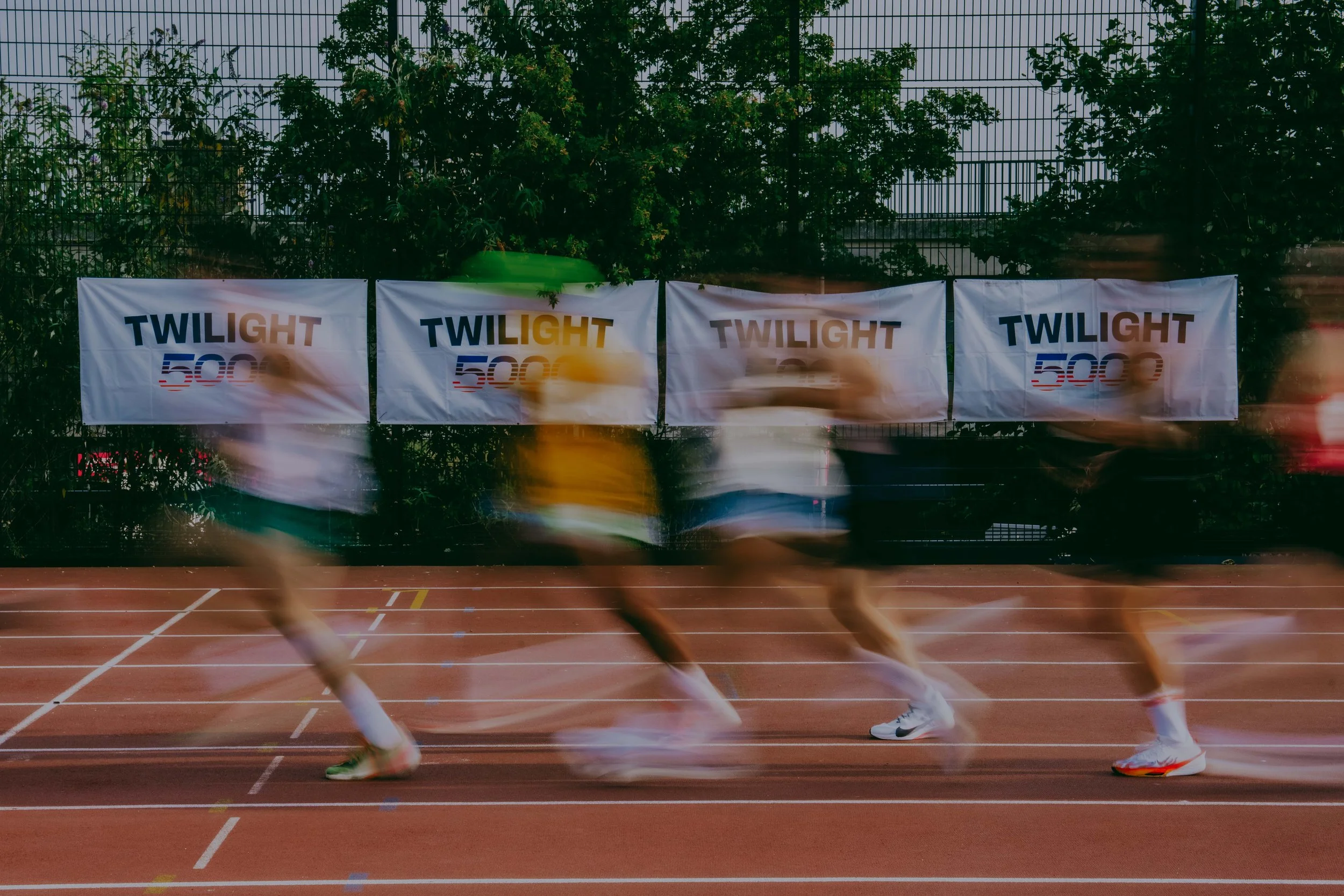 Running athletes on a track with banners reading 'Twilight 500' behind a fence surrounded by trees.