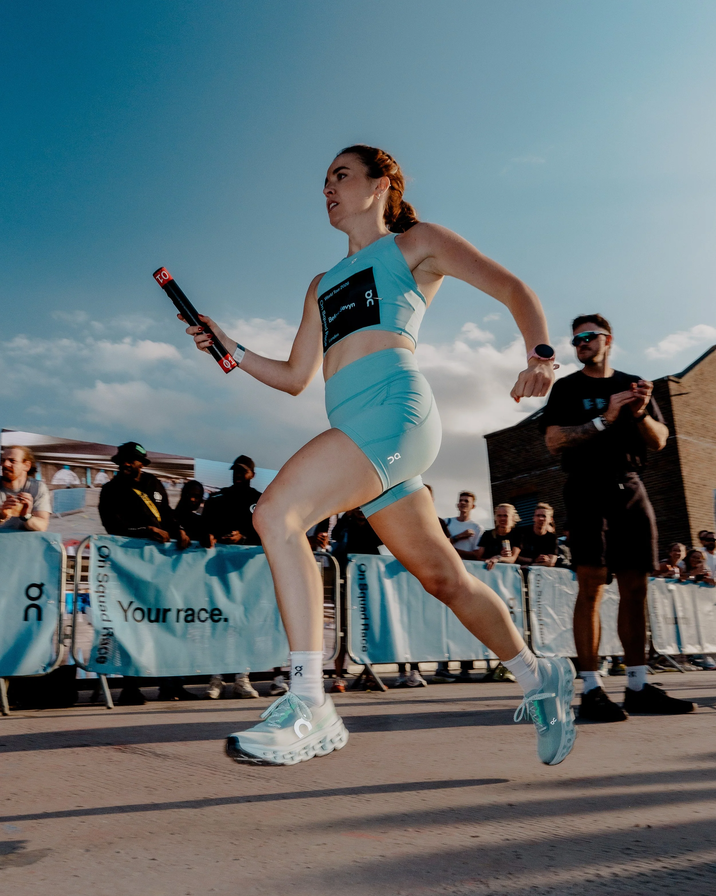 A female runner competing in a race, wearing a light blue sports outfit and running shoes, with spectators and event banners in the background on a sunny day.