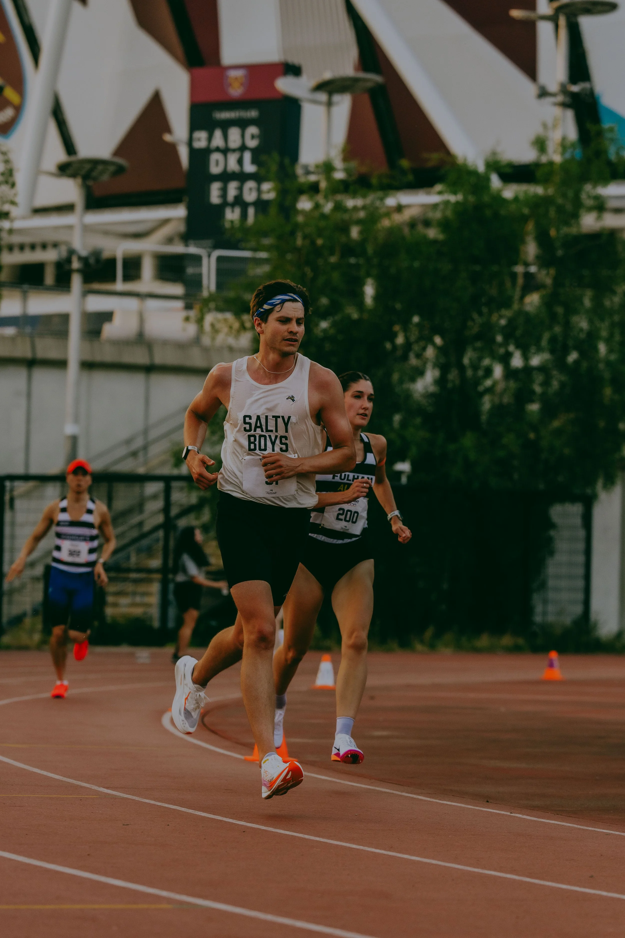 A group of women running on a track during a race, with orange cones marking the course. The woman in the foreground wears a white tank top with 