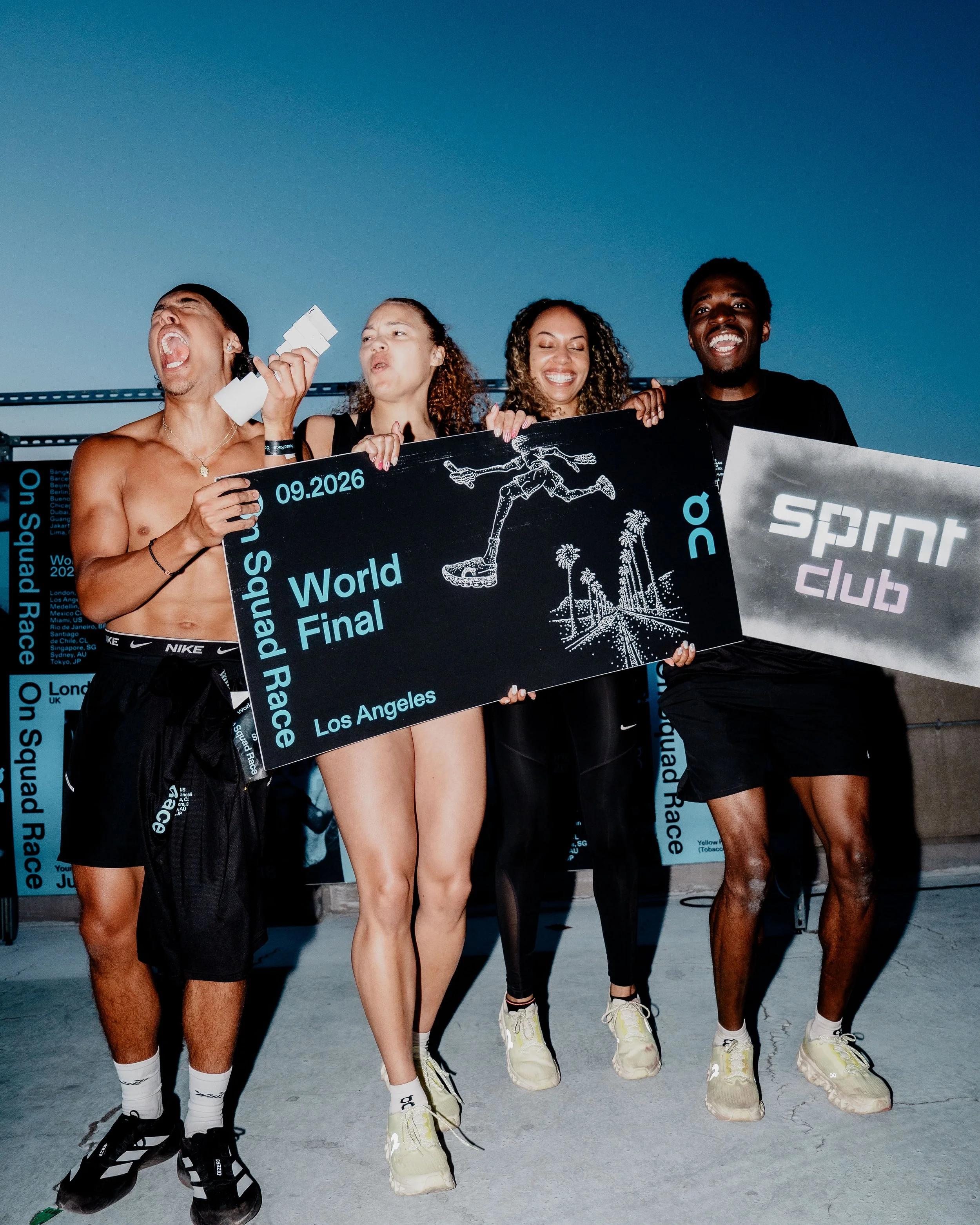 Group of four diverse young adults standing together outdoors, holding signs and smiling, at a sports or event venue during evening or night.
