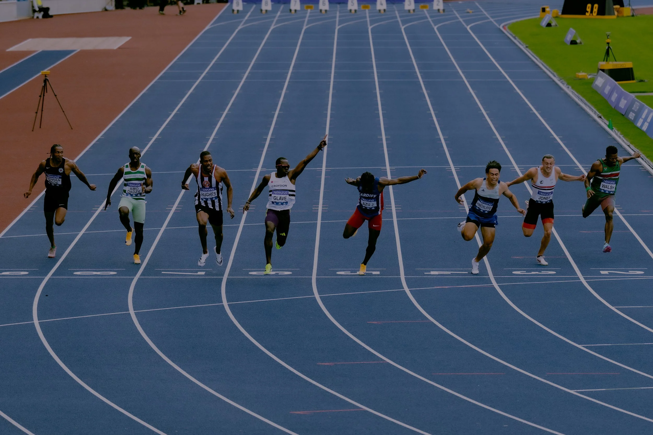 Six male sprinters racing on a blue track with white lane markings, with some athletes mid-stride and others unevenly aligned as they approach the finish line.