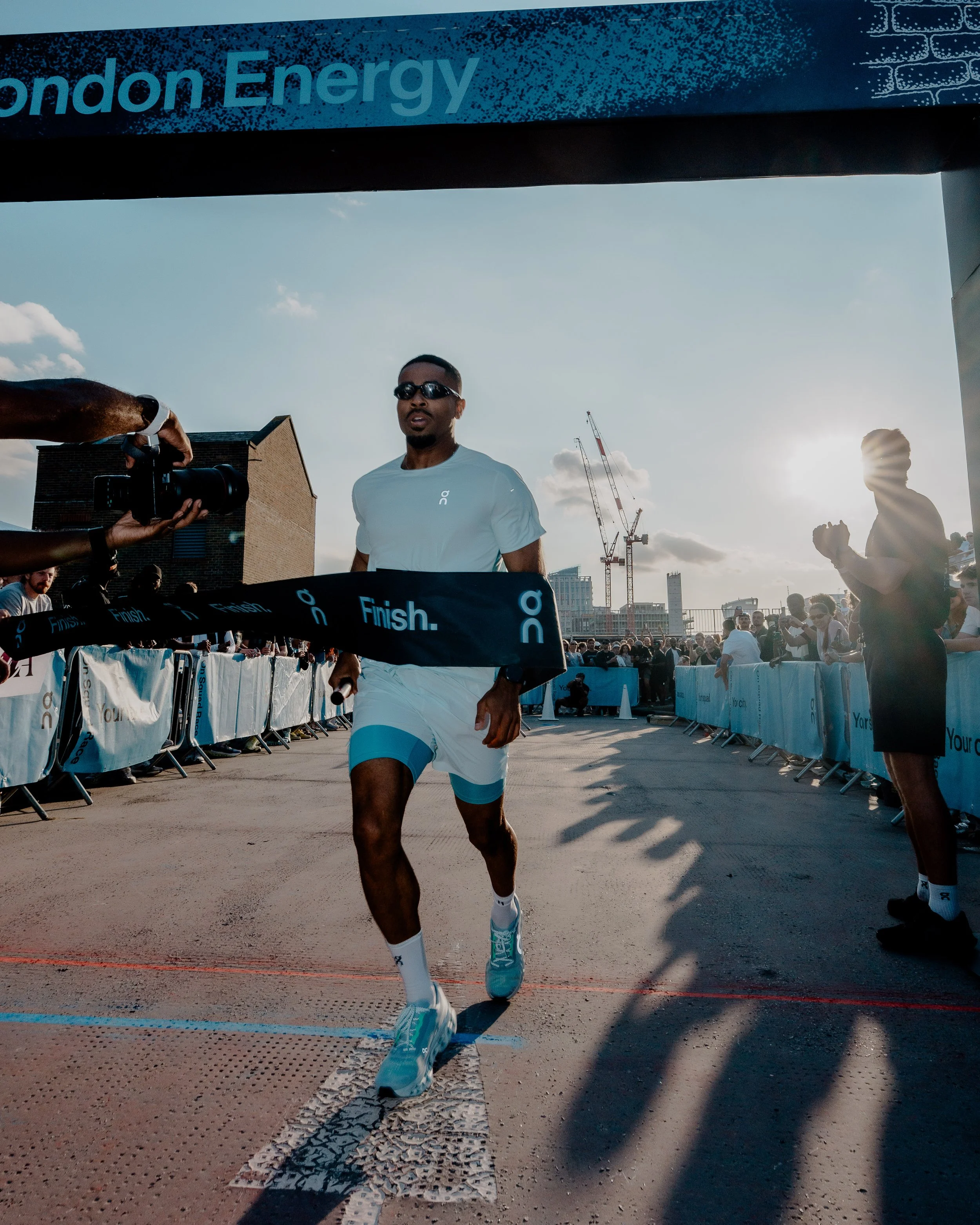 A male runner wearing sunglasses, a light blue shirt, and light blue running shoes is crossing the finish line at a race, with a crowd of spectators in the background. The time of day appears to be late afternoon or early evening.