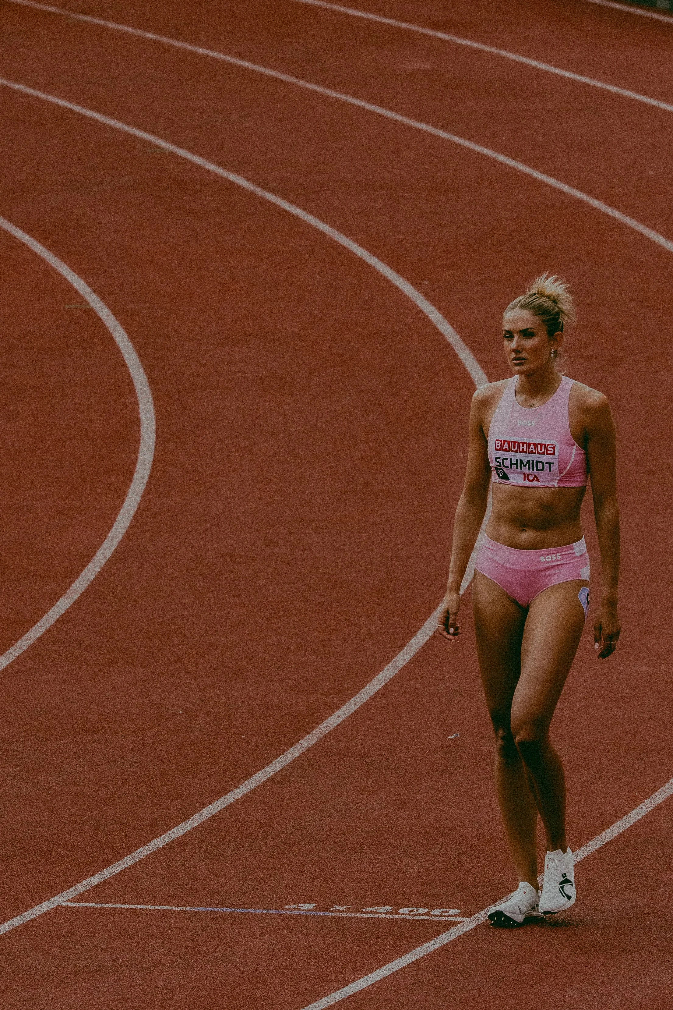 A female sprinter walking on a red running track, wearing a pink sports bra and shorts with sponsor logos, and white athletic shoes.