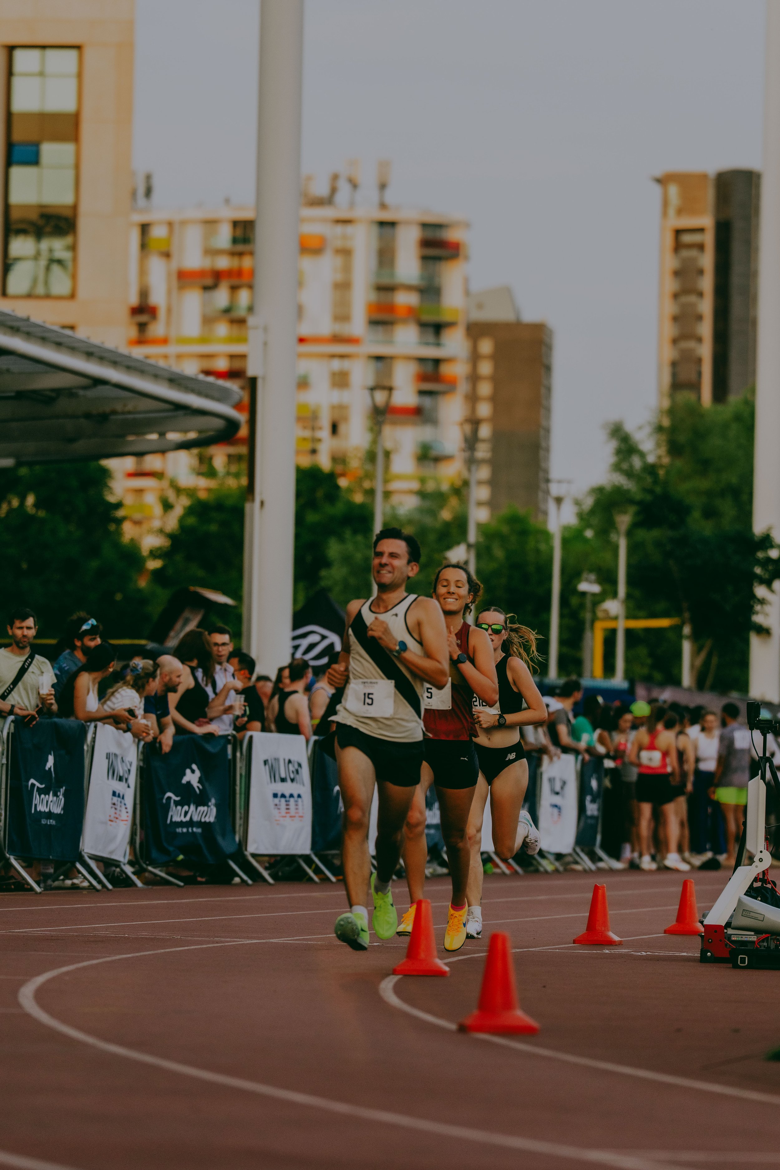 Runners participating in a race on an outdoor track, with spectators and organizational banners lining the sides, and tall buildings in the background.