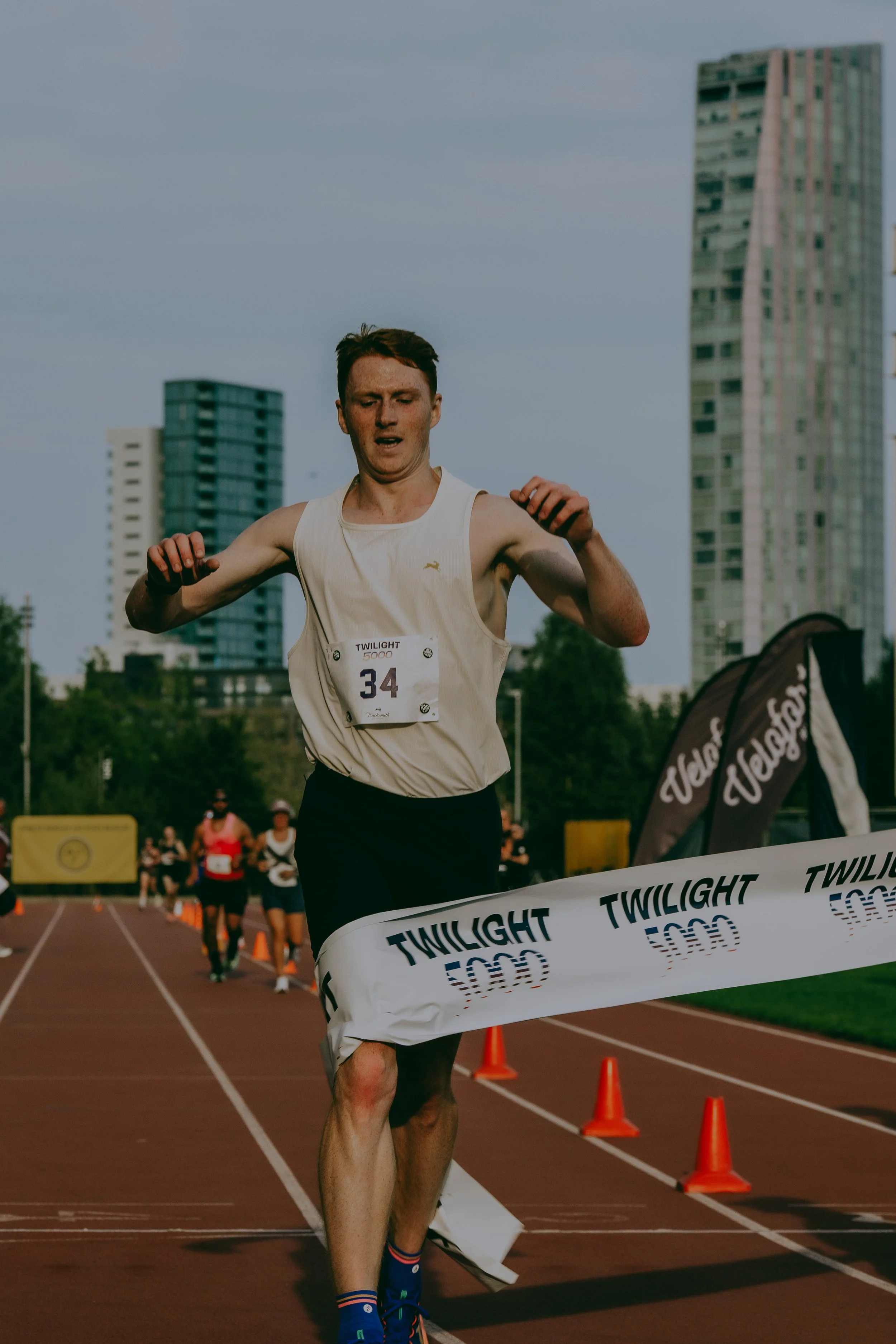 A male athlete crosses the finish line of a race on a track, with others behind him. Tall buildings are visible in the background, and orange cones mark the course. The event appears to be outdoors during daylight.