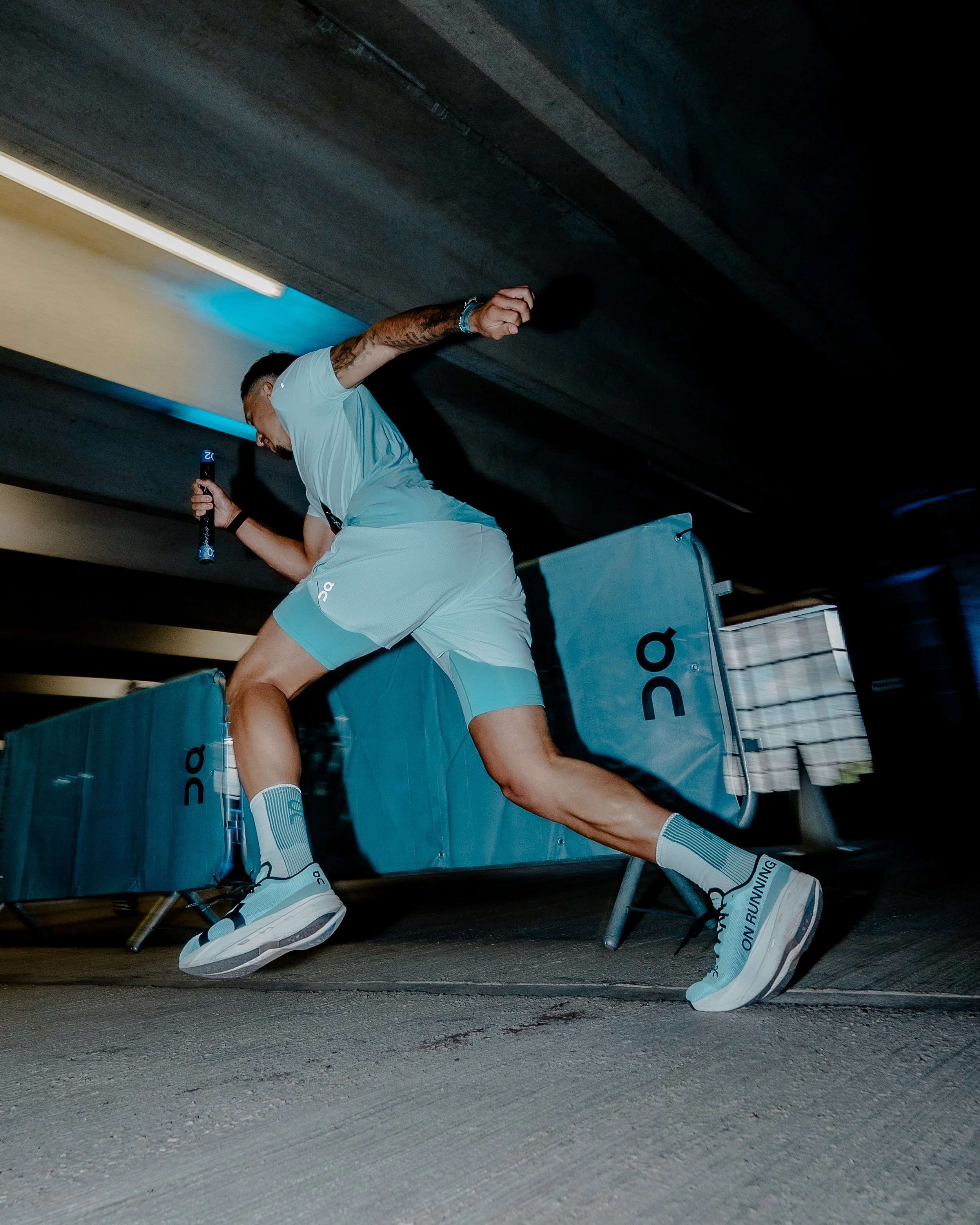 Athlete in mid-action during a race, wearing white athletic gear and holding a baton, in a sports facility with dark ceiling and bright lights.
