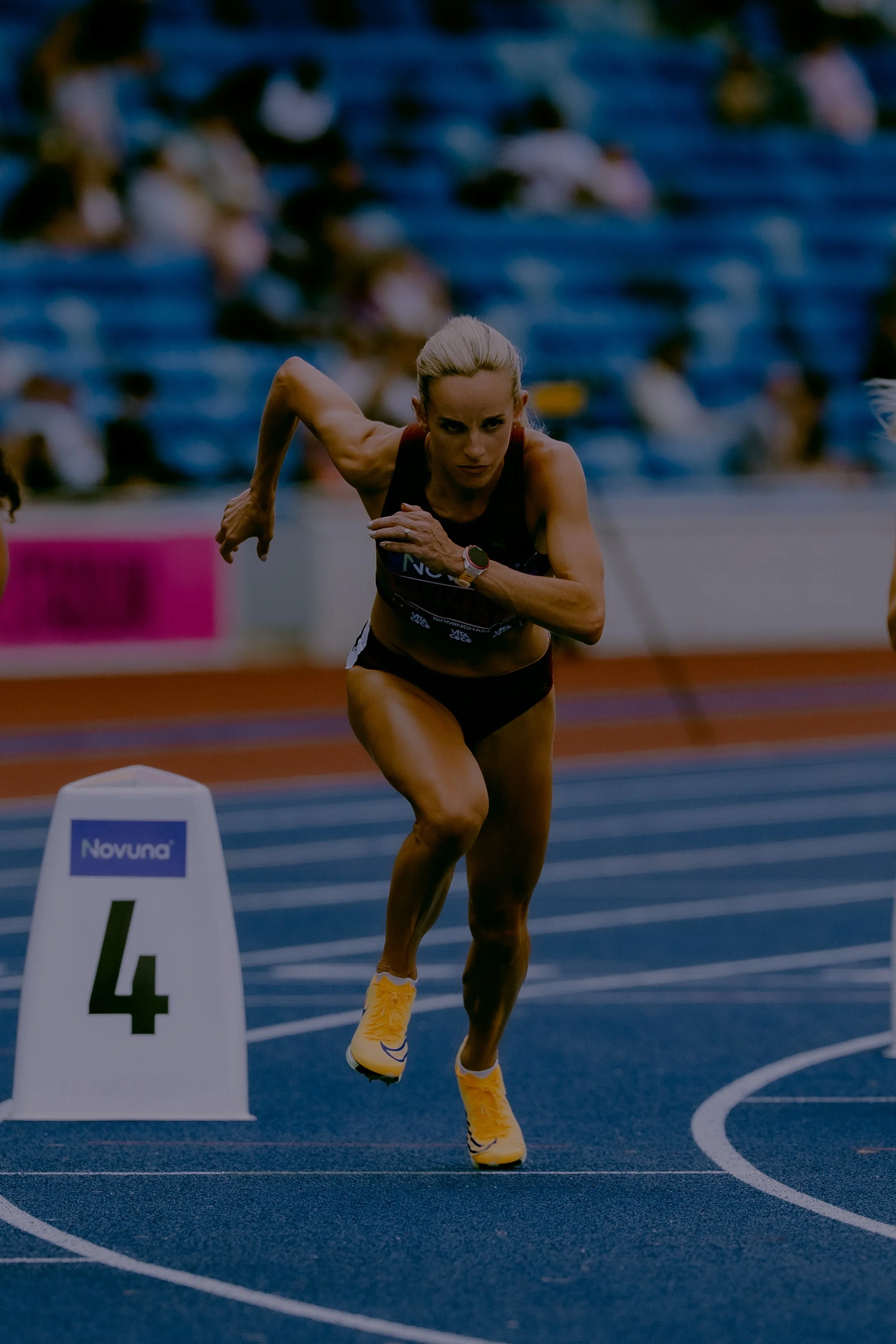 A female sprinter with blonde hair in a ponytail running on a blue track during a race, wearing yellow shoes and a dark sports outfit. The track marker reads '4' and the background shows blurred spectators.
