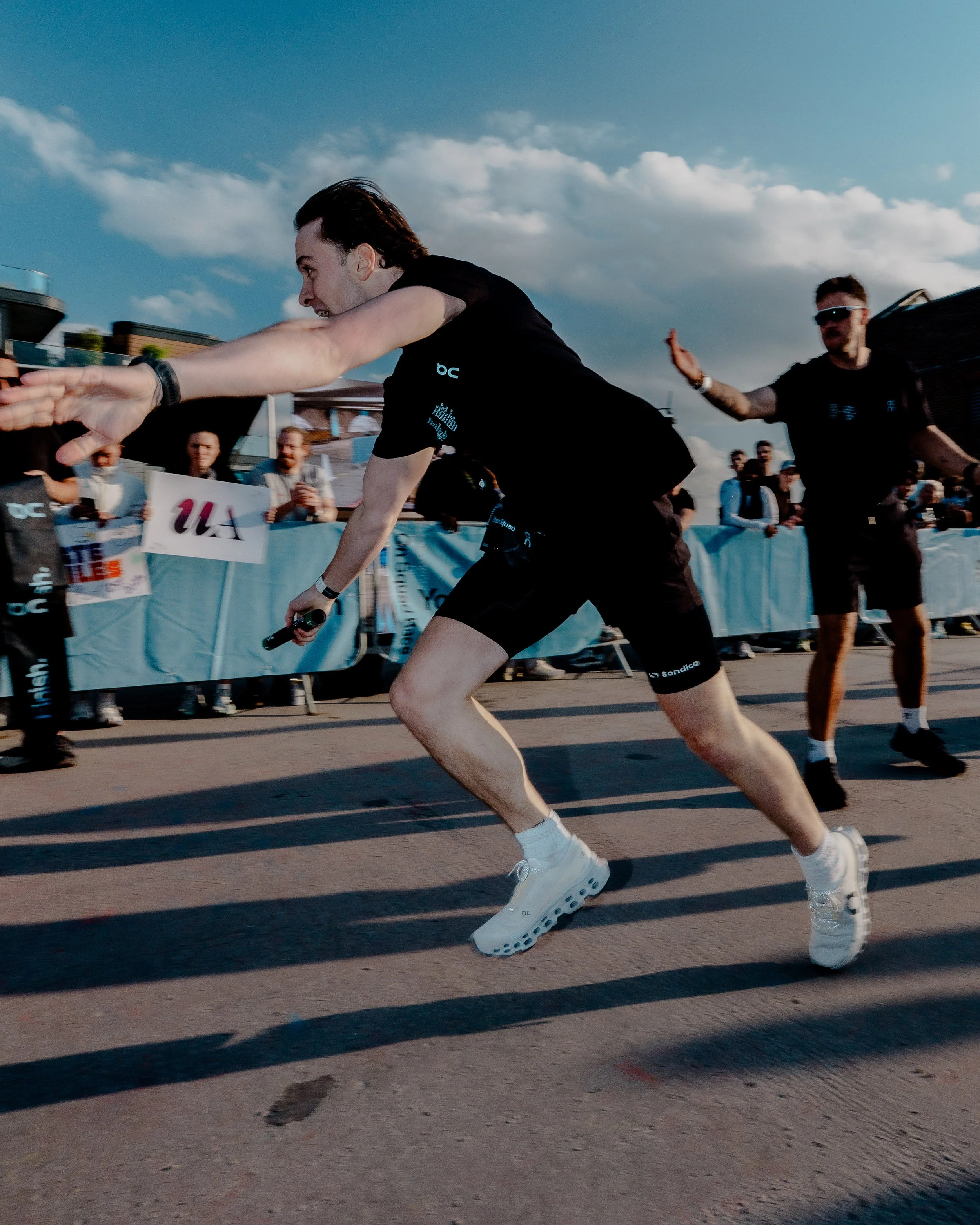 A male runner in black athletic clothes with the word 'Sondico' on his shorts running on a track during a race, with spectators behind the barrier and another man in sunglasses raising his hand.