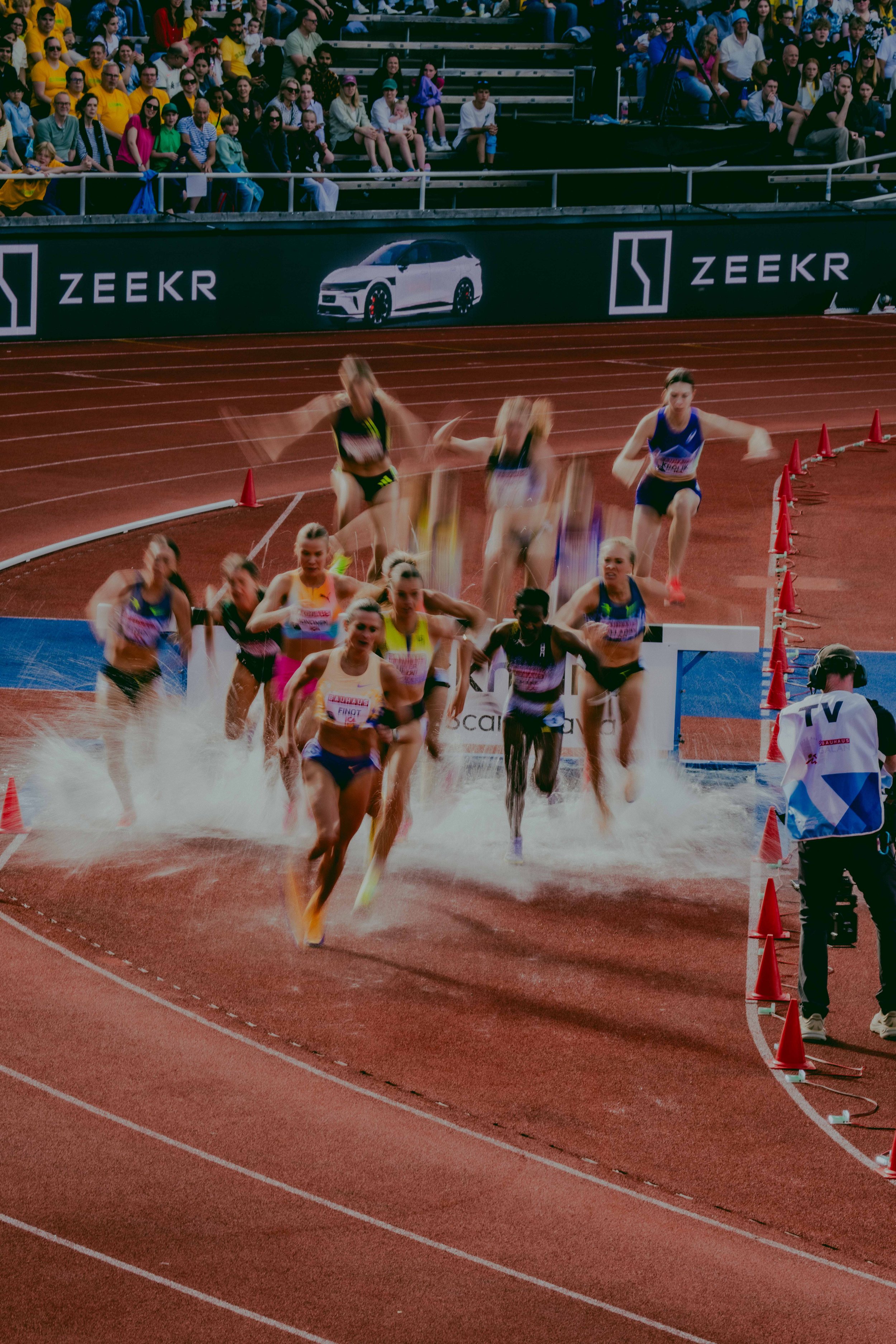 Track and field race with female athletes sprinting on a red track, spectators seated in the stands, some wearing yellow shirts, and barriers with advertising banners along the side of the track.