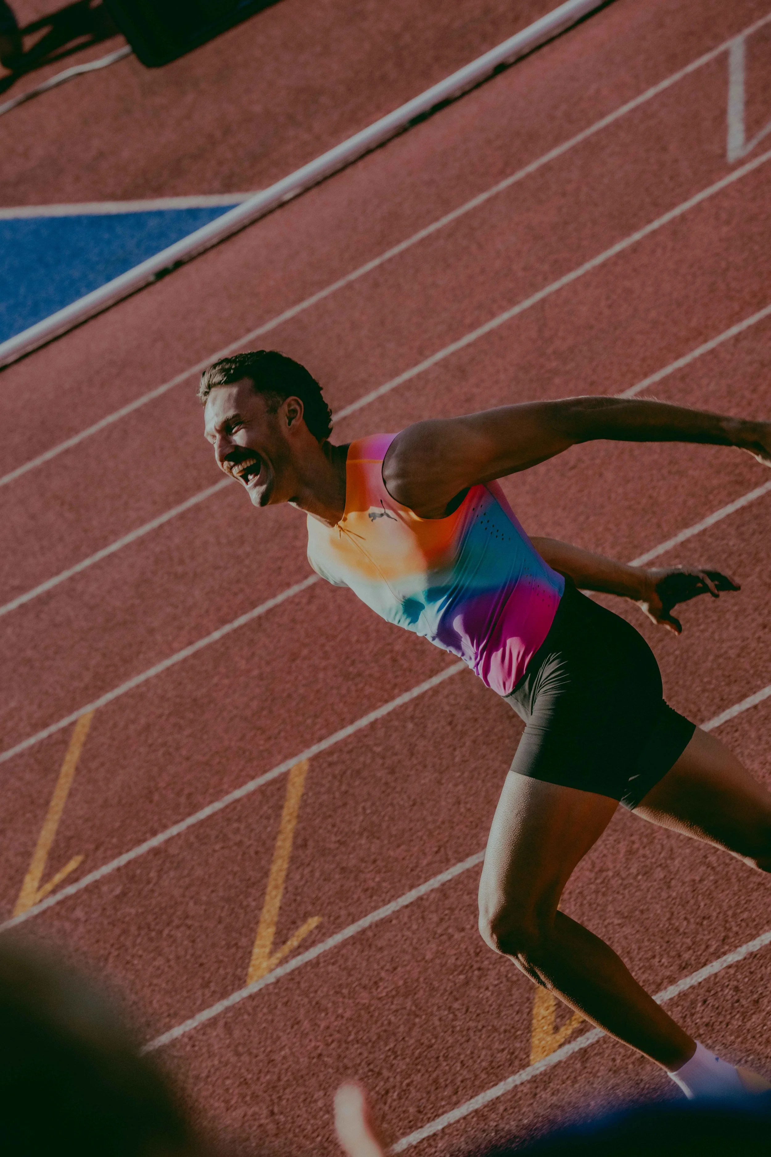 A male athlete in a colorful sports outfit is rushing on a red running track, appearing to be in the middle of a race, with an expression of effort and determination.