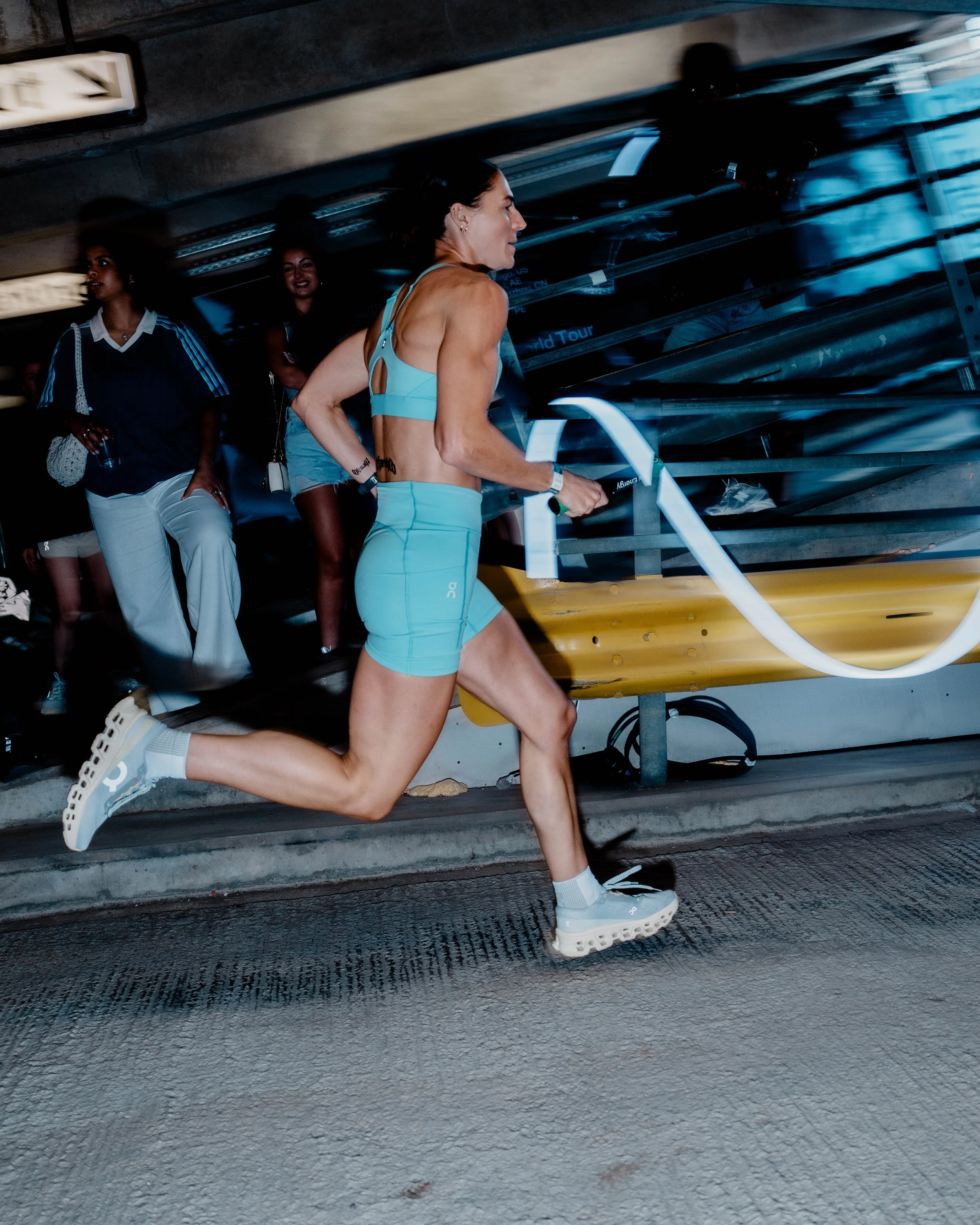 A woman jogging at night in an underground parking garage, wearing a light blue sports outfit and sneakers, with three other women walking in the background.
