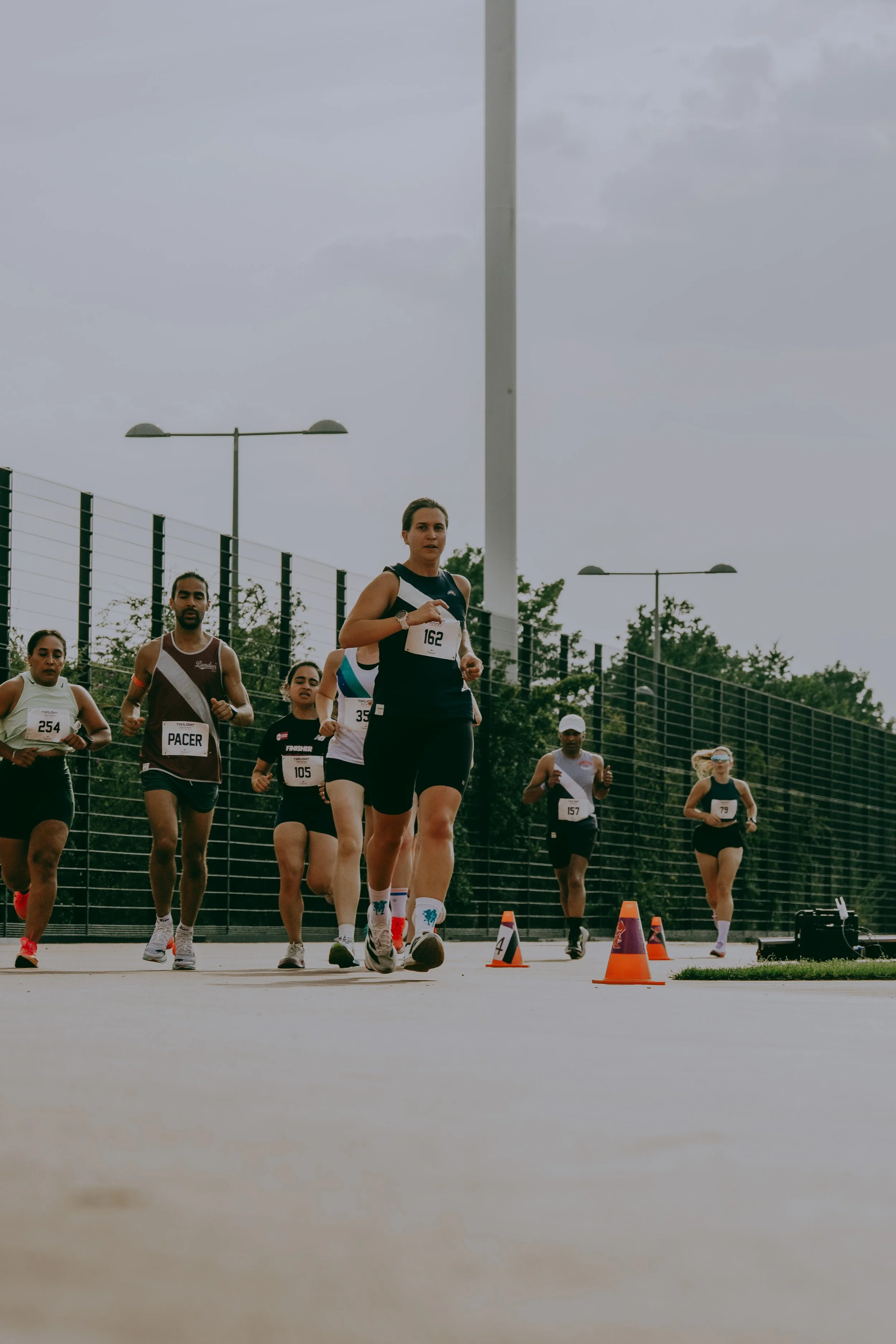 A group of runners participating in a race on a track, with some wearing bibs displaying race numbers, orange cones marking the course, and a fence with trees and streetlights in the background on a cloudy day.