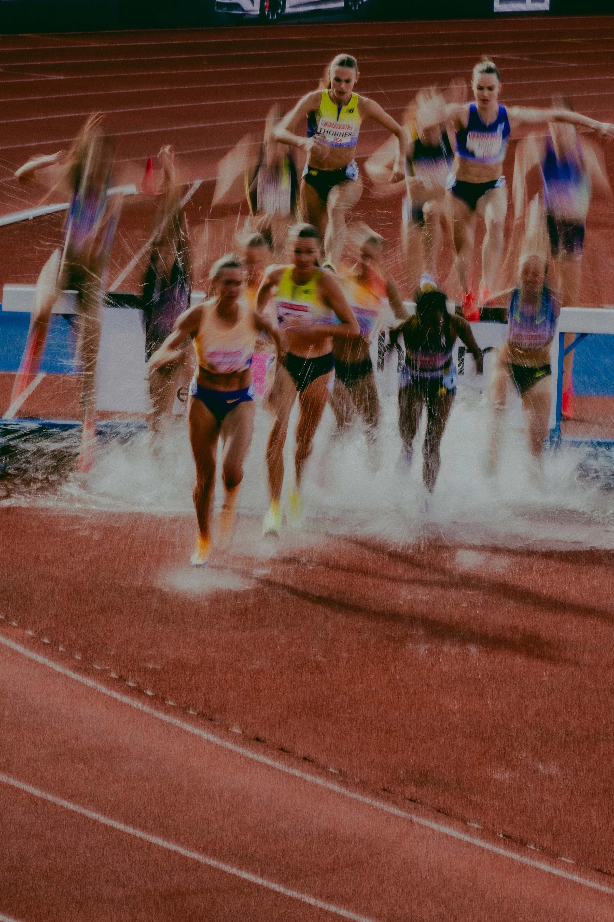 Track and field athletes running out of water on a red running track after a race, with some athletes drenched in water, in an outdoor stadium.