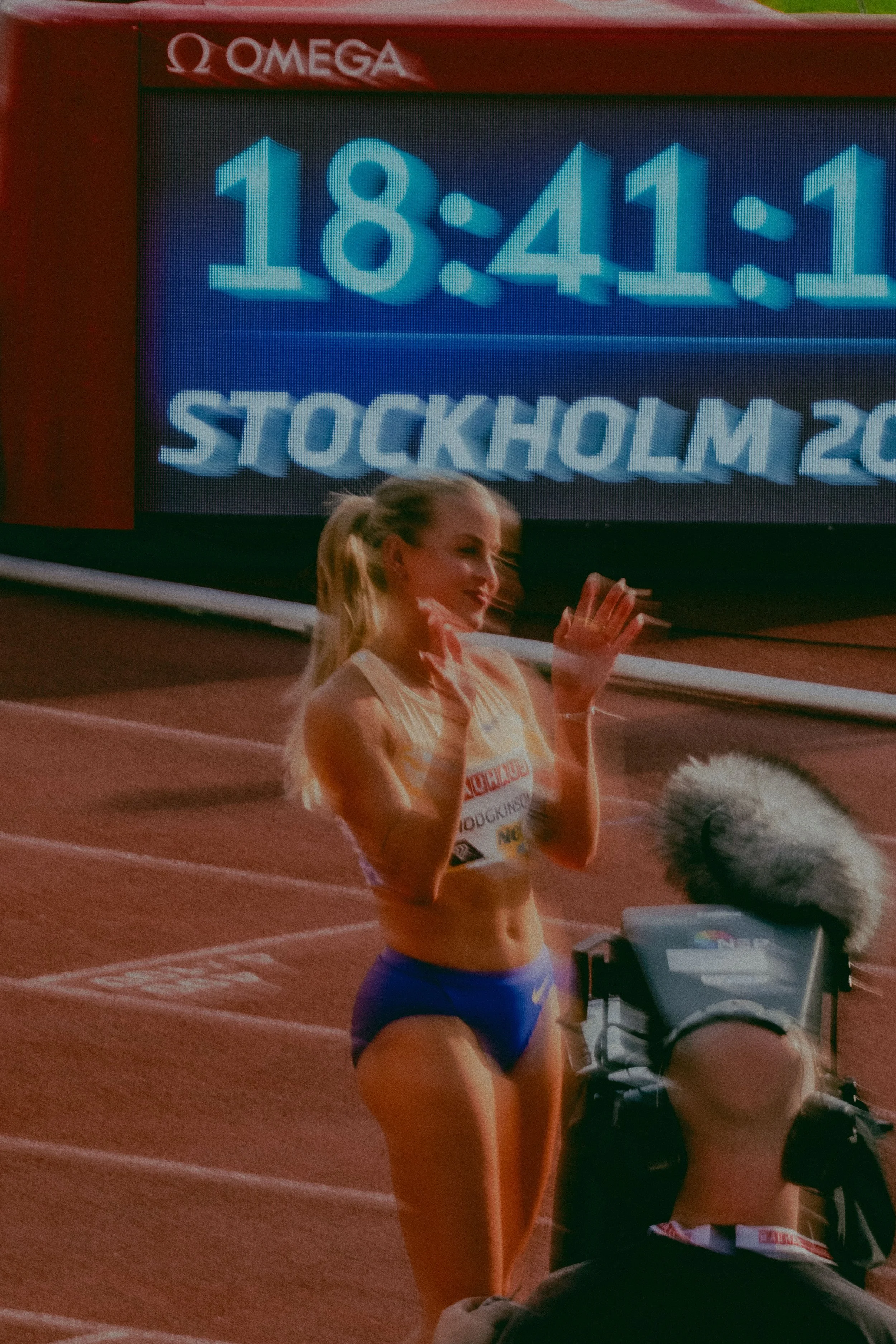 A female athlete in athletic attire standing on a running track at the Stockholm 2022 event, with a digital clock displaying 18:41:11 and a partially visible person in athletic gear.