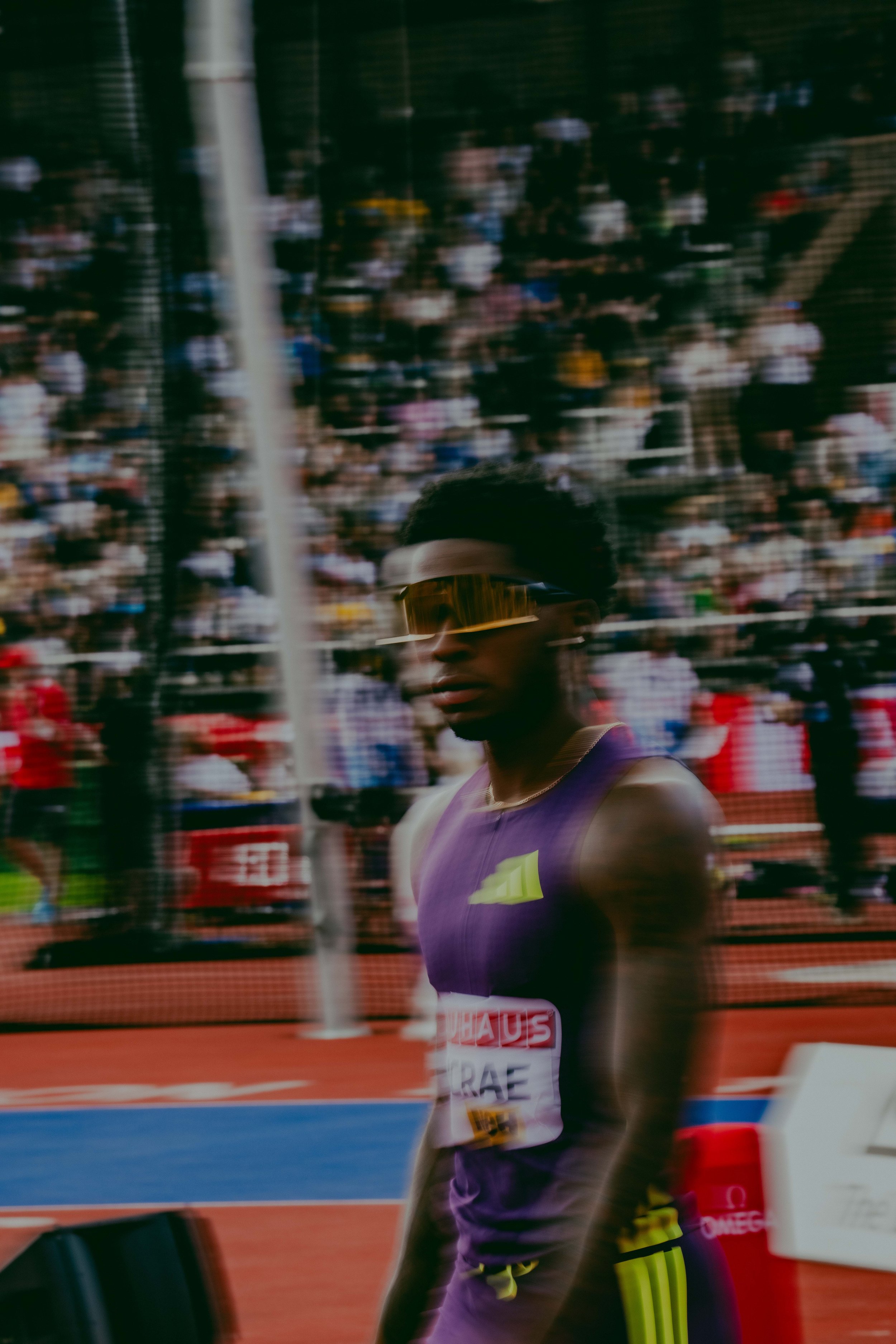 A male athlete wearing a purple running uniform and sunglasses in a stadium during a race.