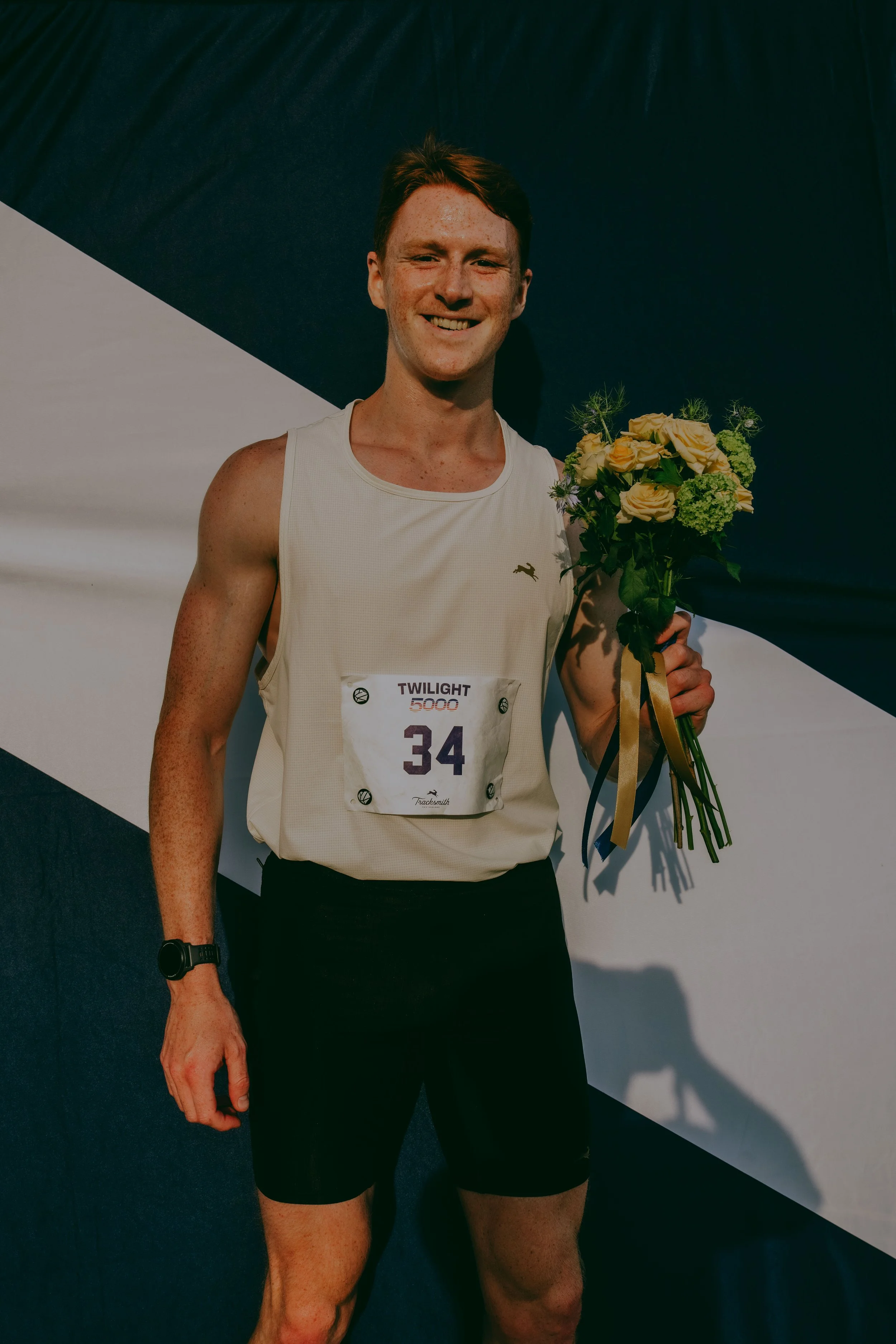 A young man with red hair and freckles holding a bouquet of flowers, smiling, wearing a white sleeveless athletic shirt with a bib number 34, standing against a black and white background.