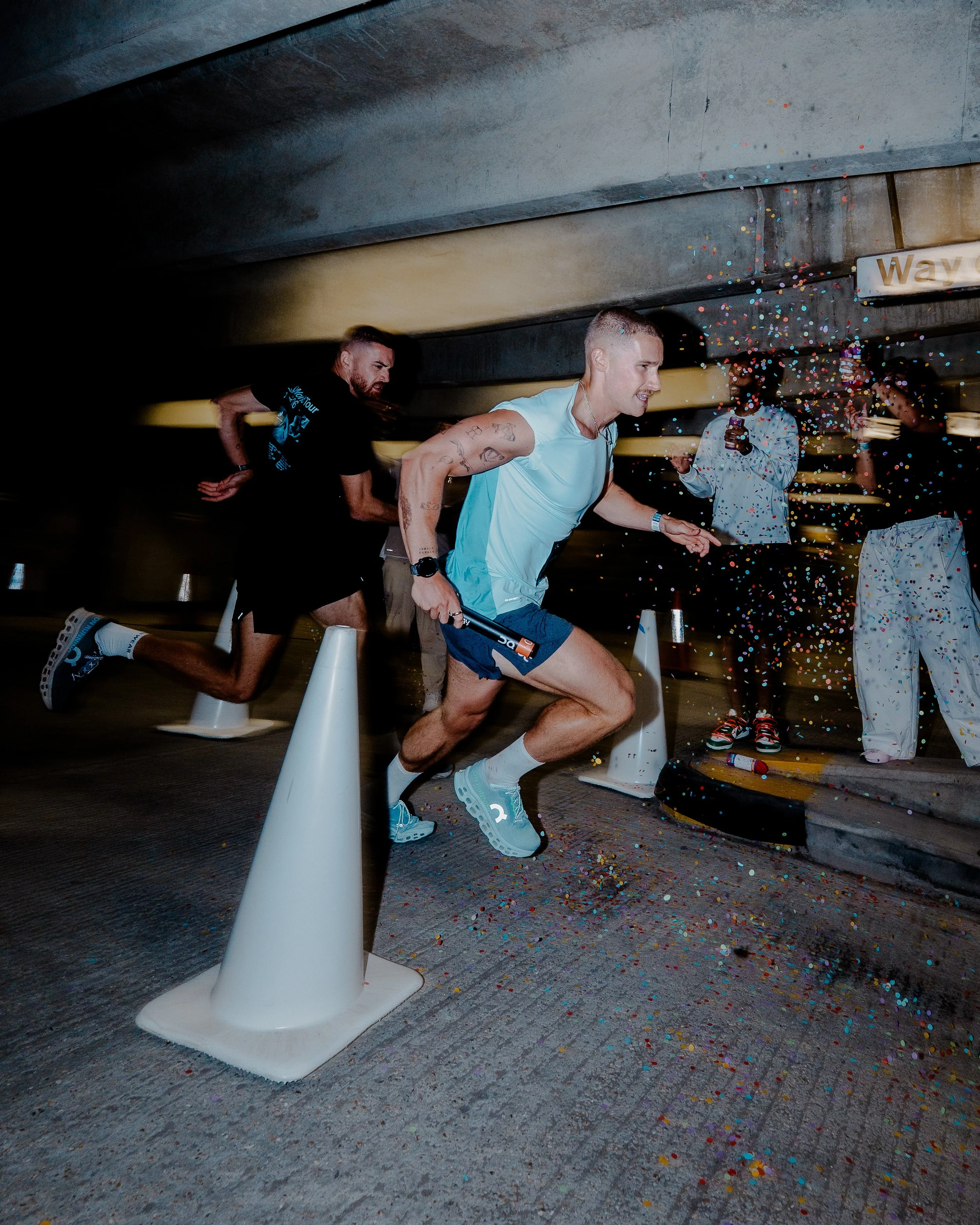 People celebrating under a concrete structure, running past cones with confetti falling around them, some holding phones, with a sign that partially reads 'Way' in the background.