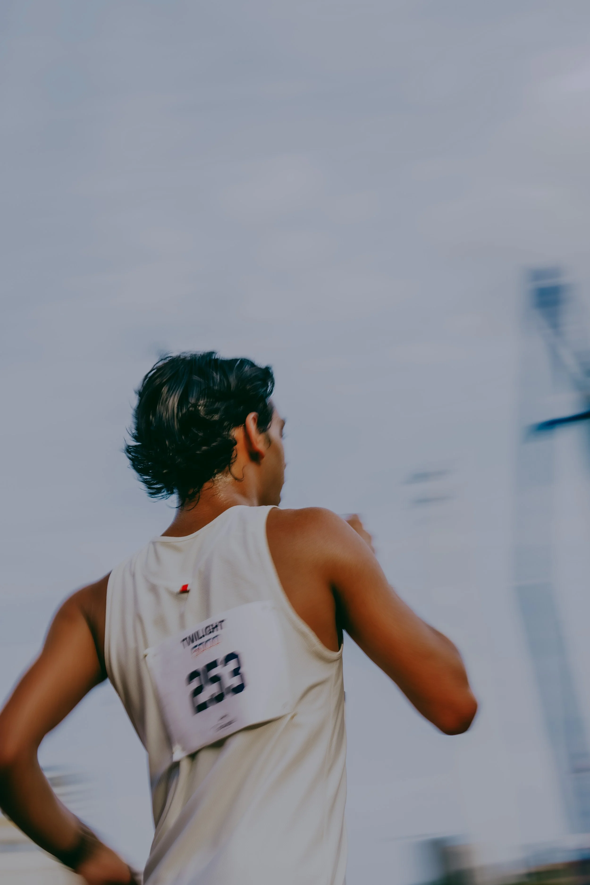 A person with short dark hair, wearing a white sleeveless athletic shirt with a bib number, running outdoors. The sky in the background is cloudy.