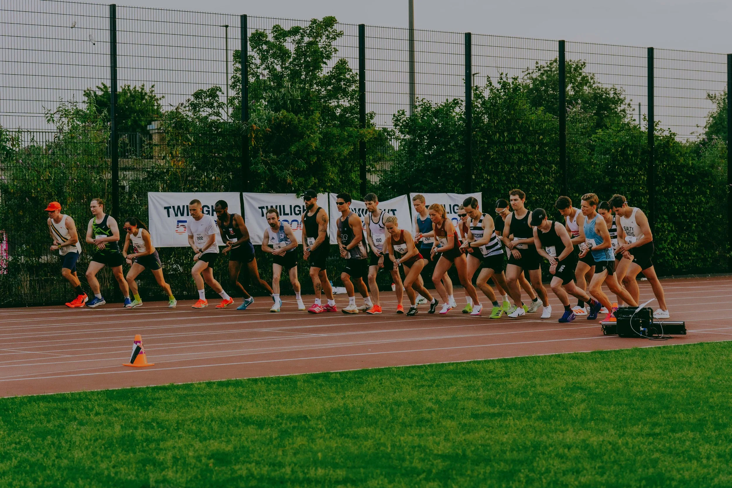 A group of runners at the start of a race on a track, with trees and a tall fence in the background, and orange cones marking the lanes.