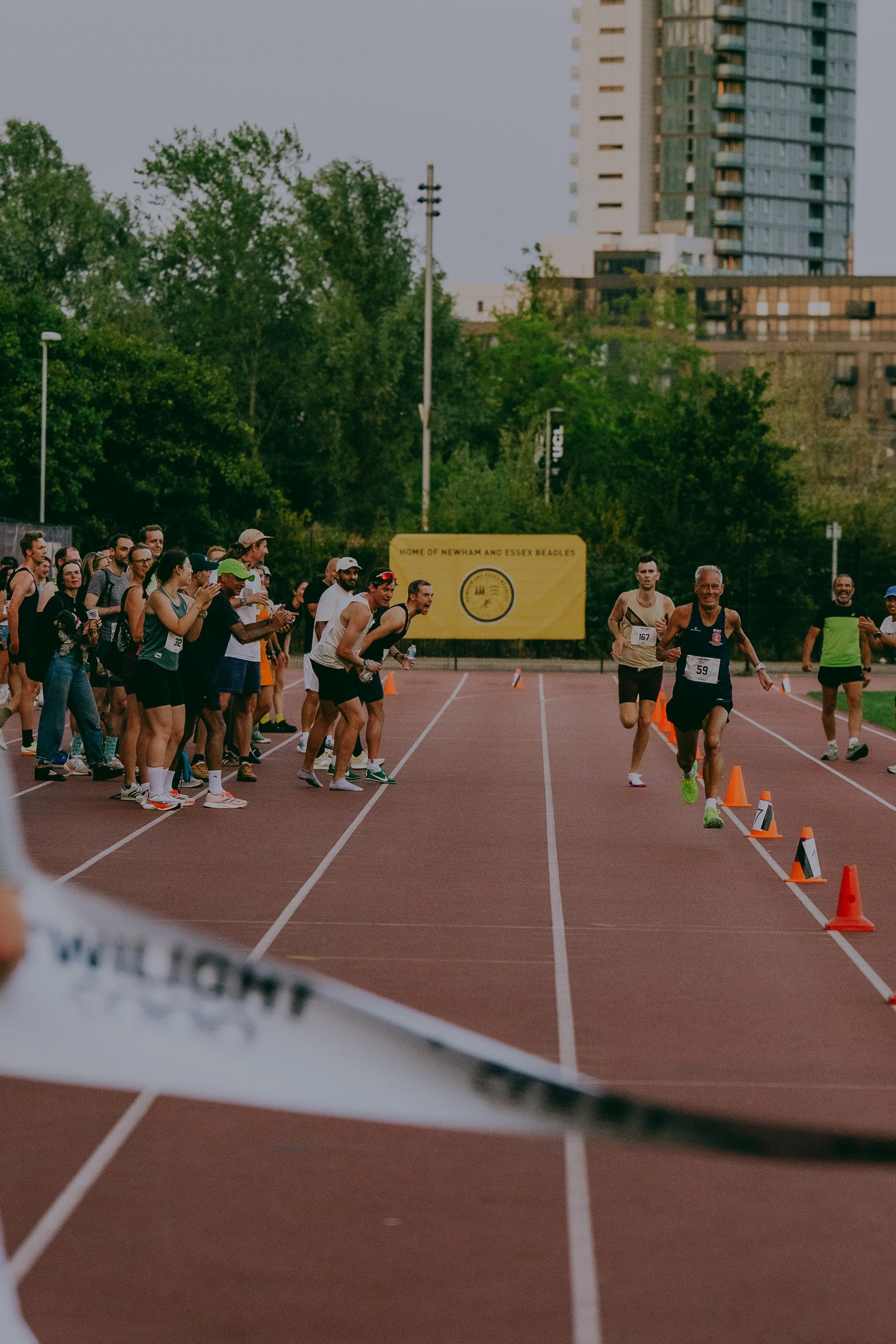 A group of runners competing in a race on an outdoor track, with spectators on the sidelines and a large yellow banner in the background. The scene is set in an urban area with high-rise buildings visible in the distance.
