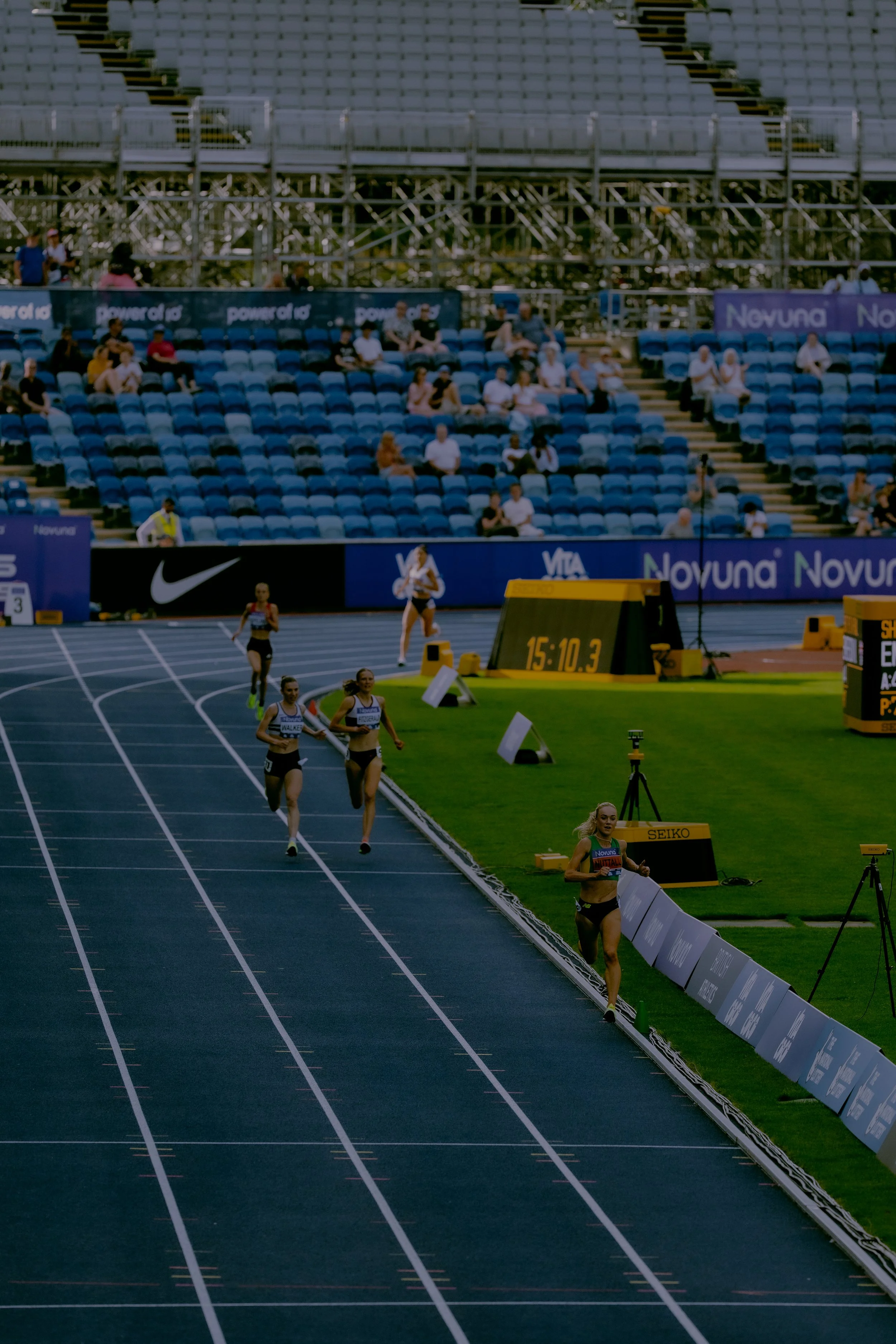 Athletes competing in a track and field race at an outdoor stadium, with spectators seated in the bleachers and digital timers visible.