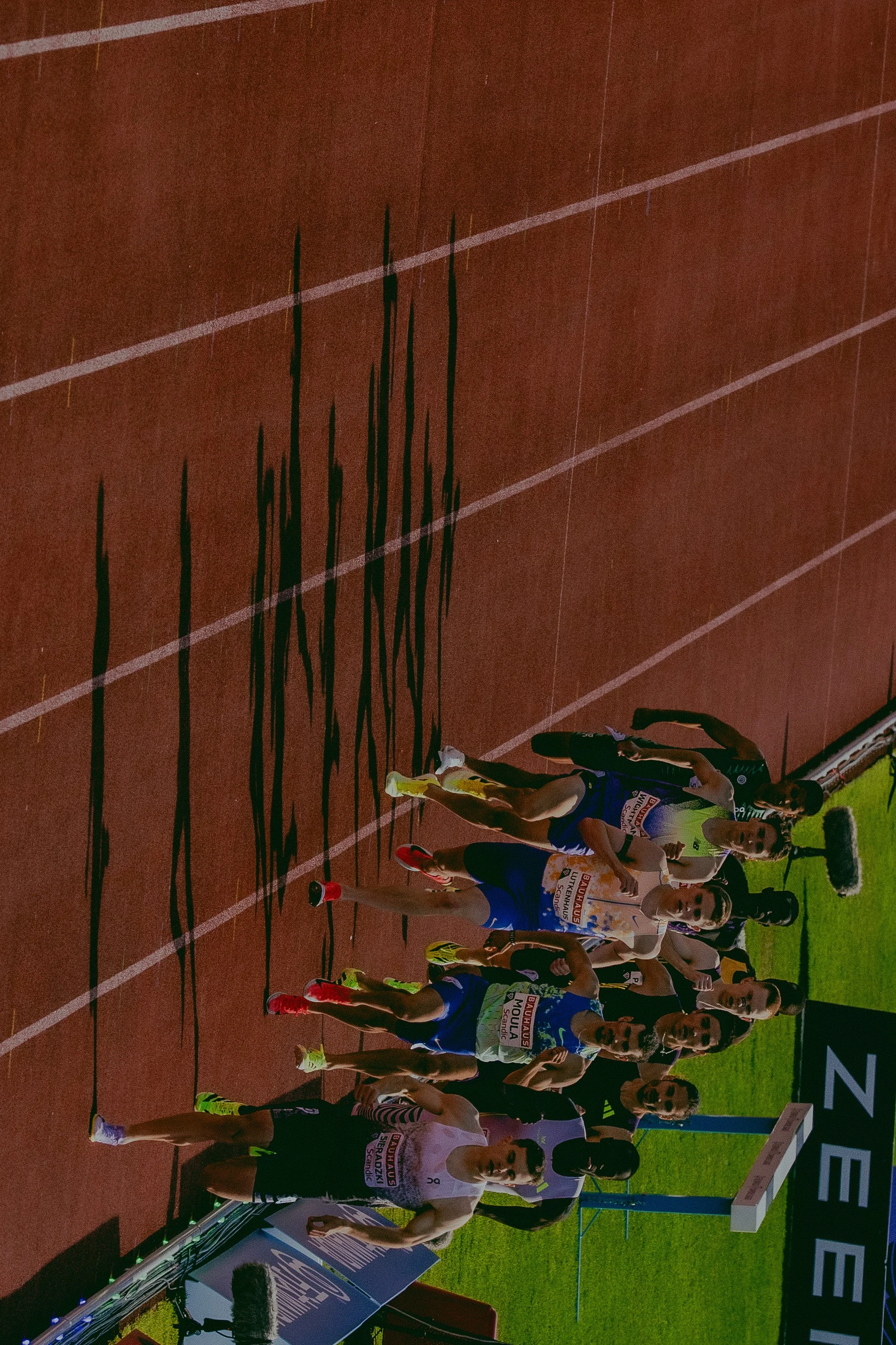 Athletes standing on a running track at an outdoor track and field event, prepared for a race, with some athletes wearing running uniforms and sports gear.