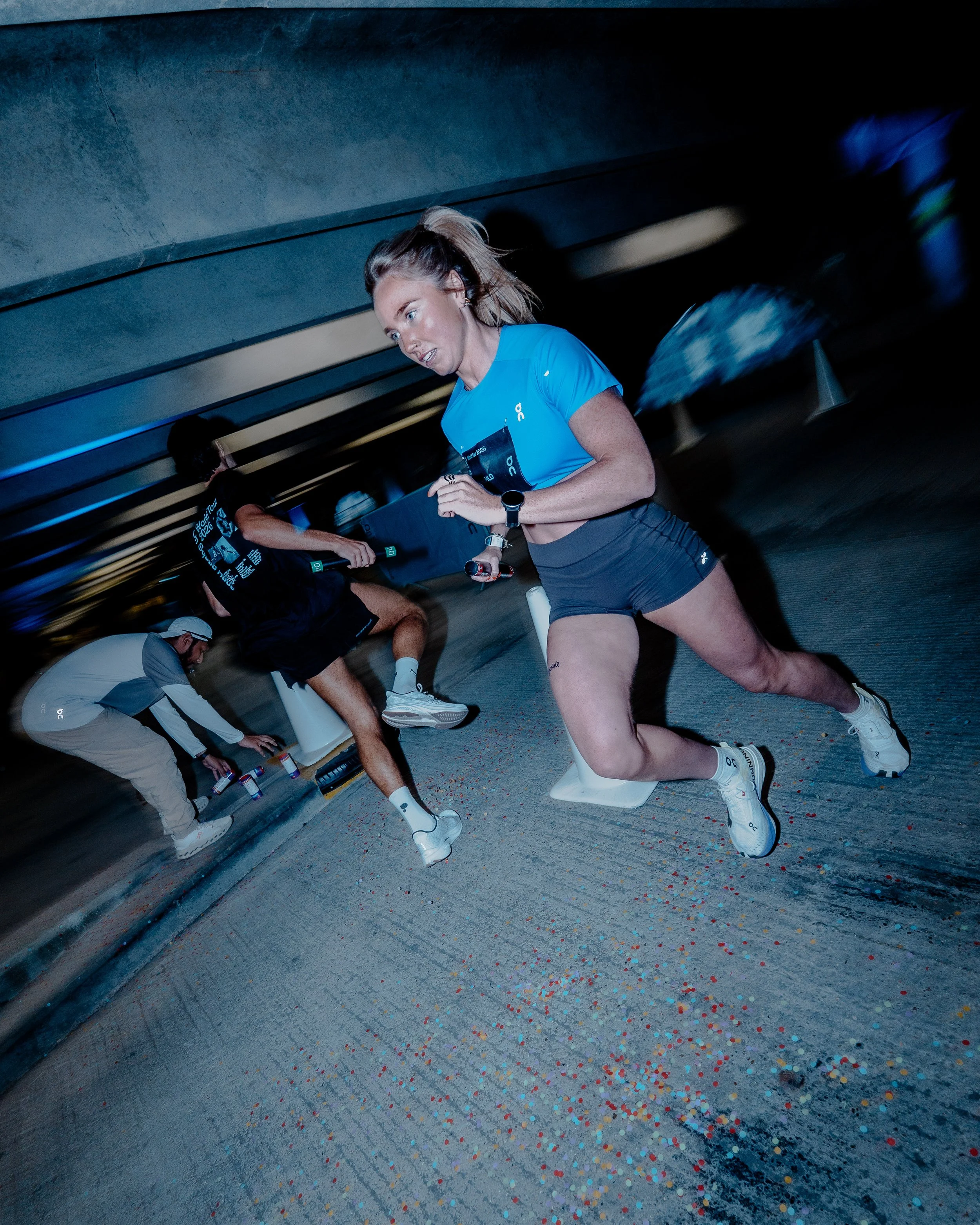 A woman in athletic clothing running in an indoor setting with colorful confetti on the floor. She has a focused expression and is wearing a blue shirt, black shorts, white shoes, and a watch. There are other people and a person kneeling down visible