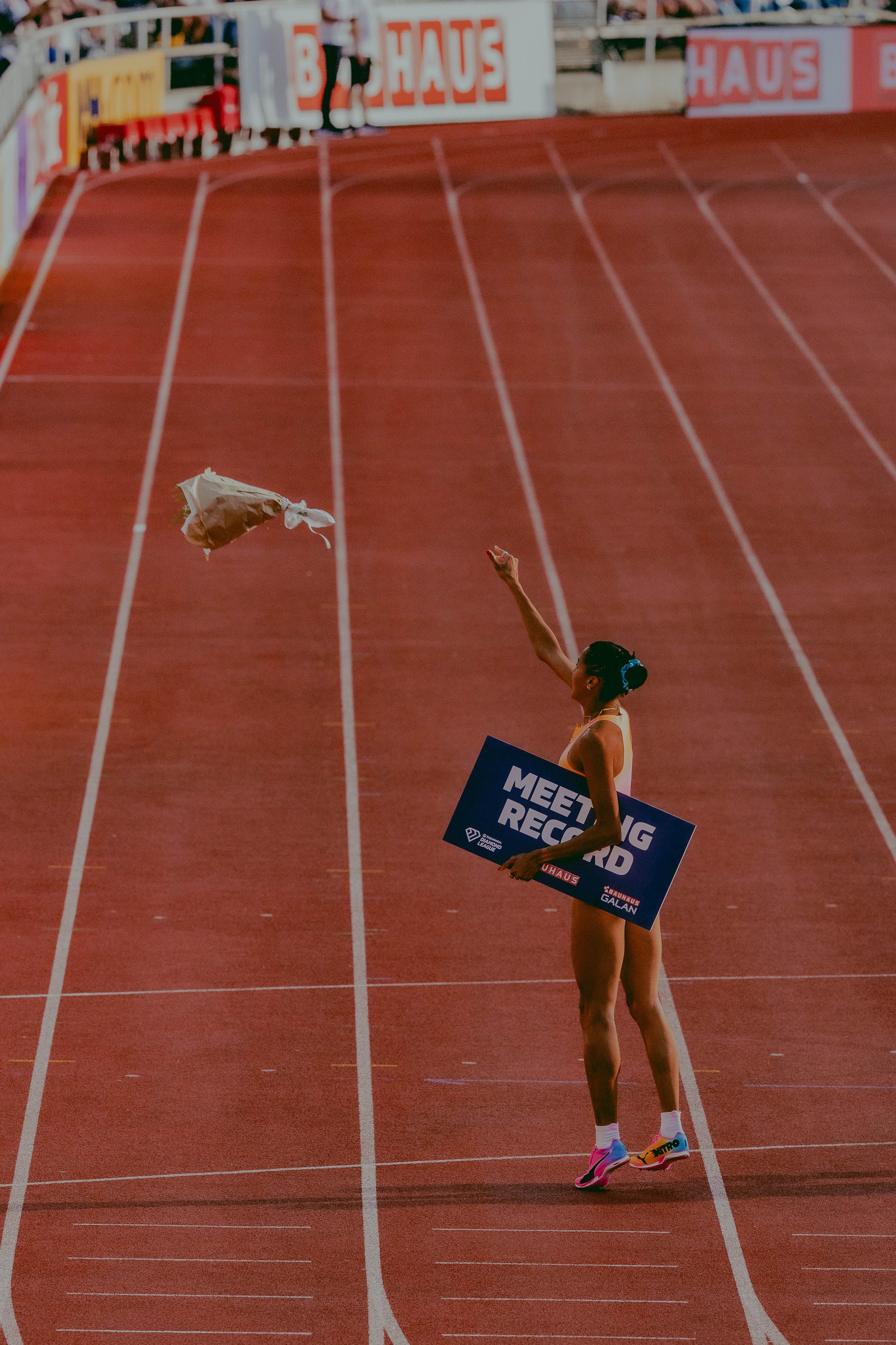 A female athlete in a track stadium holding a sign that says 'MEETING RECORD'; she is standing on the track, wearing colorful running shoes, shorts, and a sleeveless outfit, with her hair tied back in a bun. A person is seen in the background near th