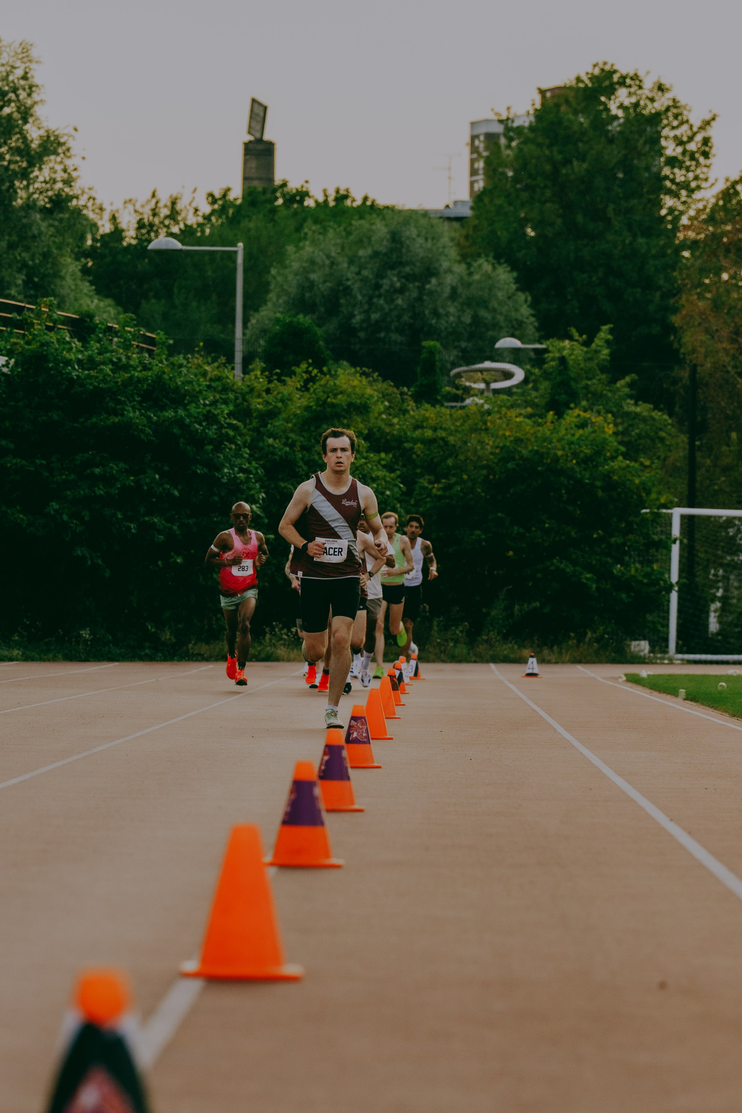 People running in a race on an outdoor track, with orange cones marking the lanes and trees in the background during sunset.