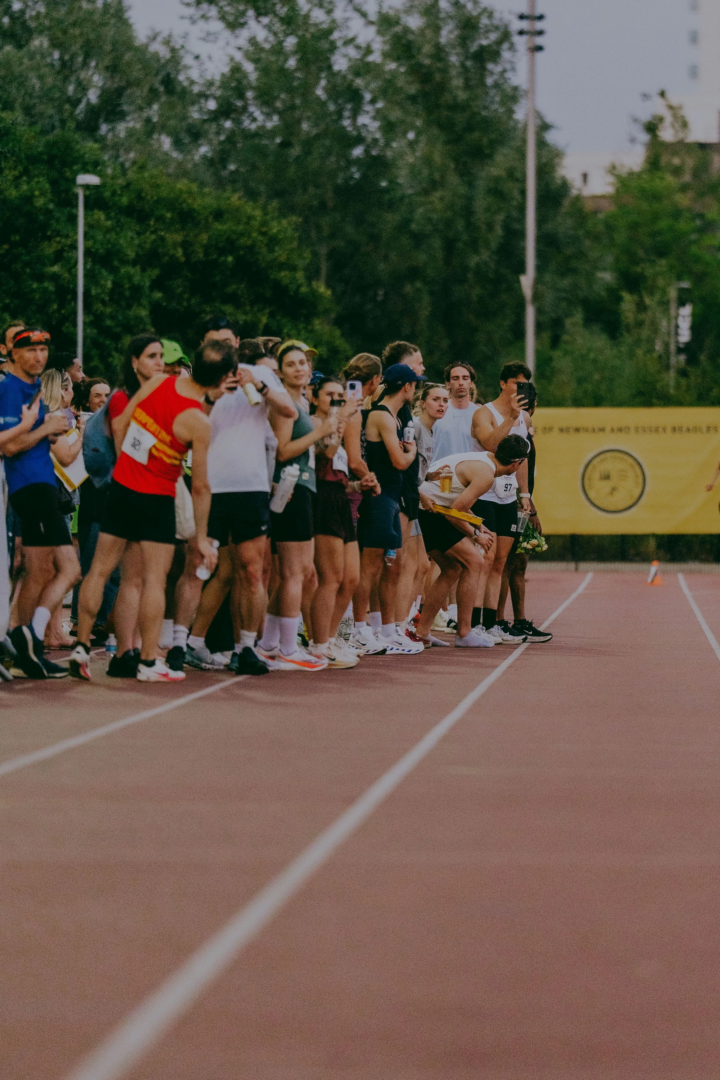 Group of runners at the starting line of a track race, some holding drinks and others adjusting their gear, with a yellow banner and green trees in the background.