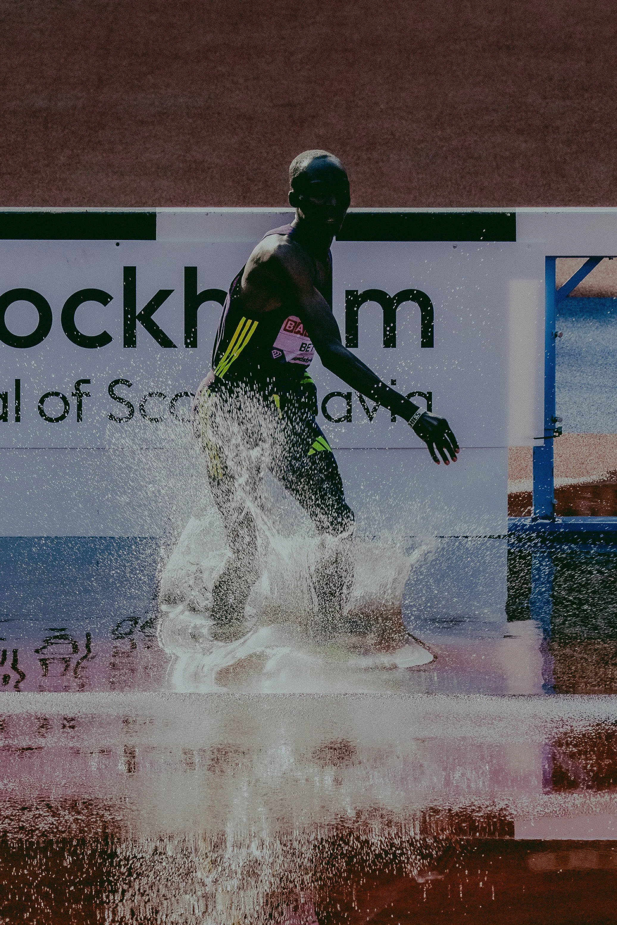 A male athlete running through water on a track, with water splashing around his legs, at an outdoor sports event.