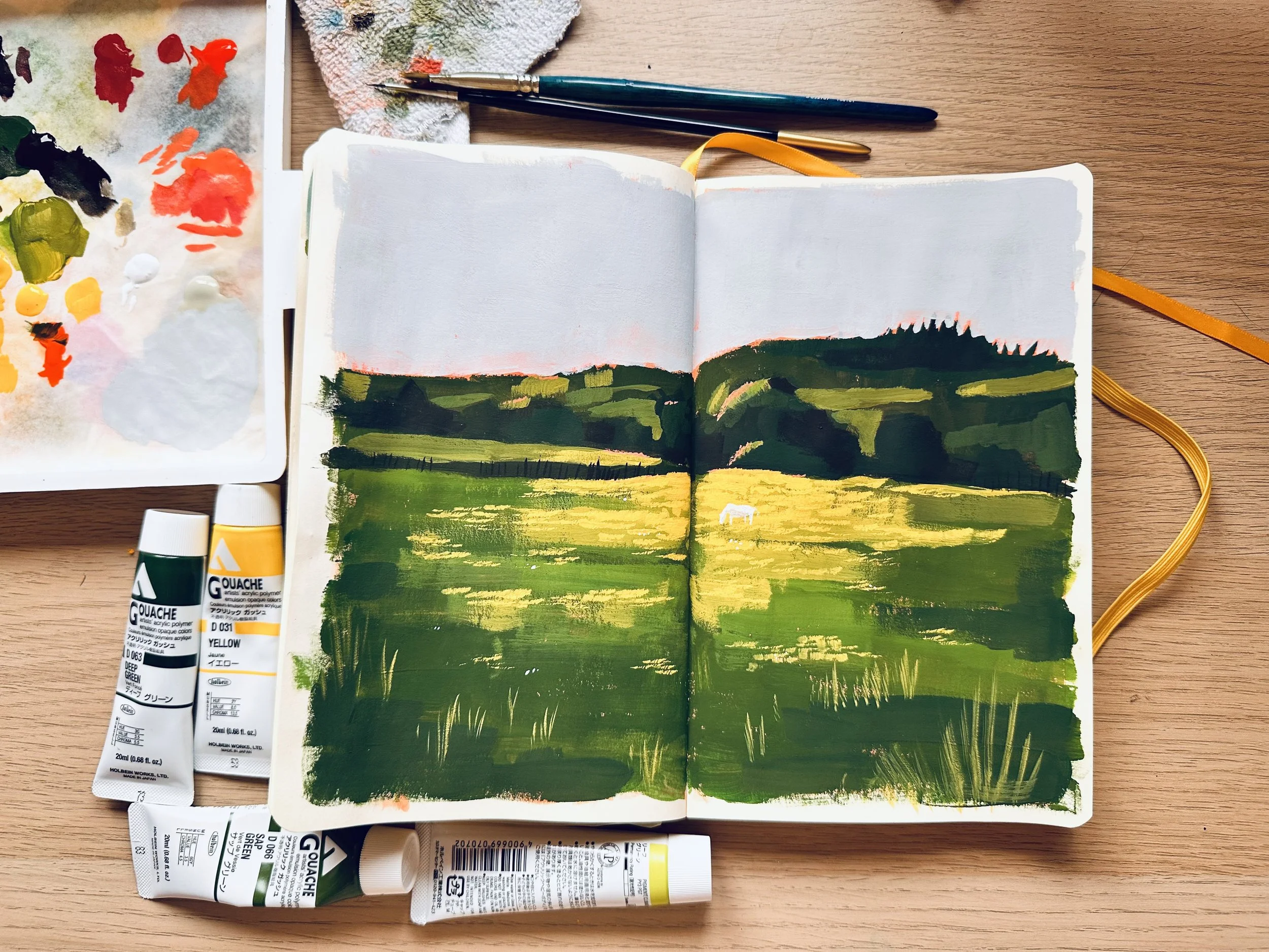 An open sketchbook with a landscape painting of green fields, yellow grass, and distant dark hills, surrounded by watercolor paints and brushes on a wooden table.