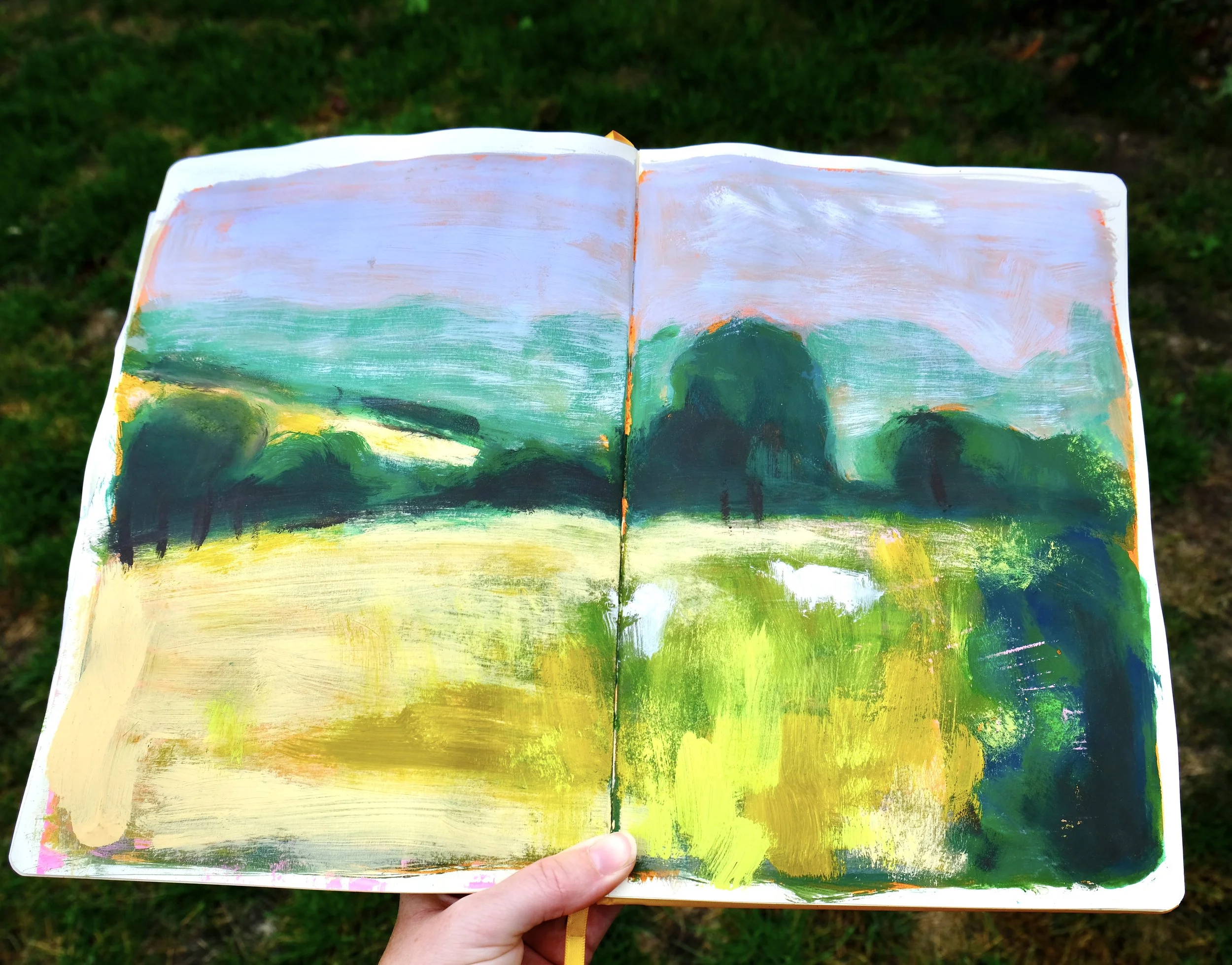 An open sketchbook with a colorful landscape painting, featuring trees, mountains, sky, and grass, held outdoors against a grassy background.