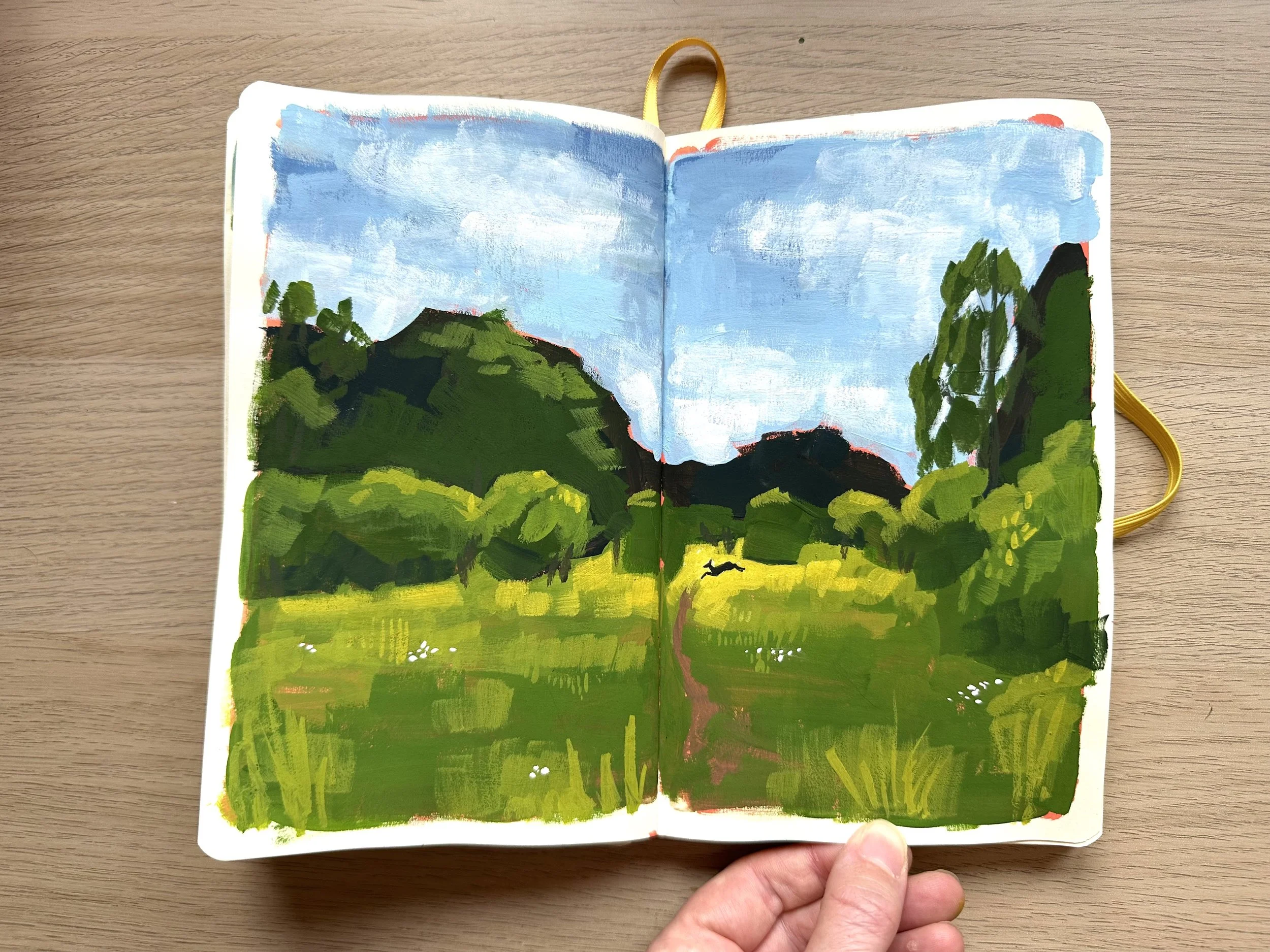 A hand holding an open sketchbook with a painting of a green landscape, trees, mountains, and a blue sky.