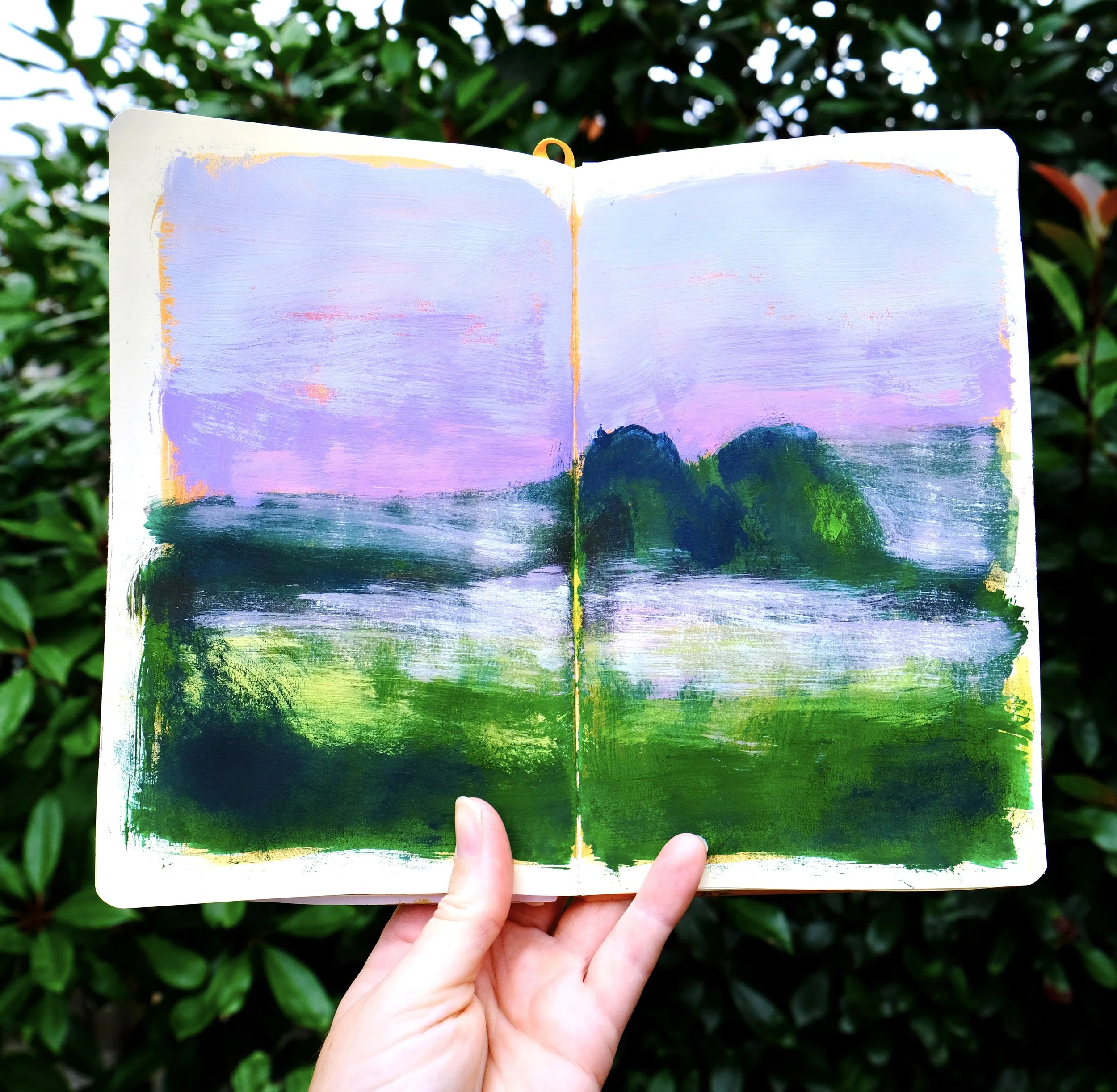 A hand holding an open sketchbook with a colorful landscape painting of a sky, mountains, and green fields. Green foliage is in the background.