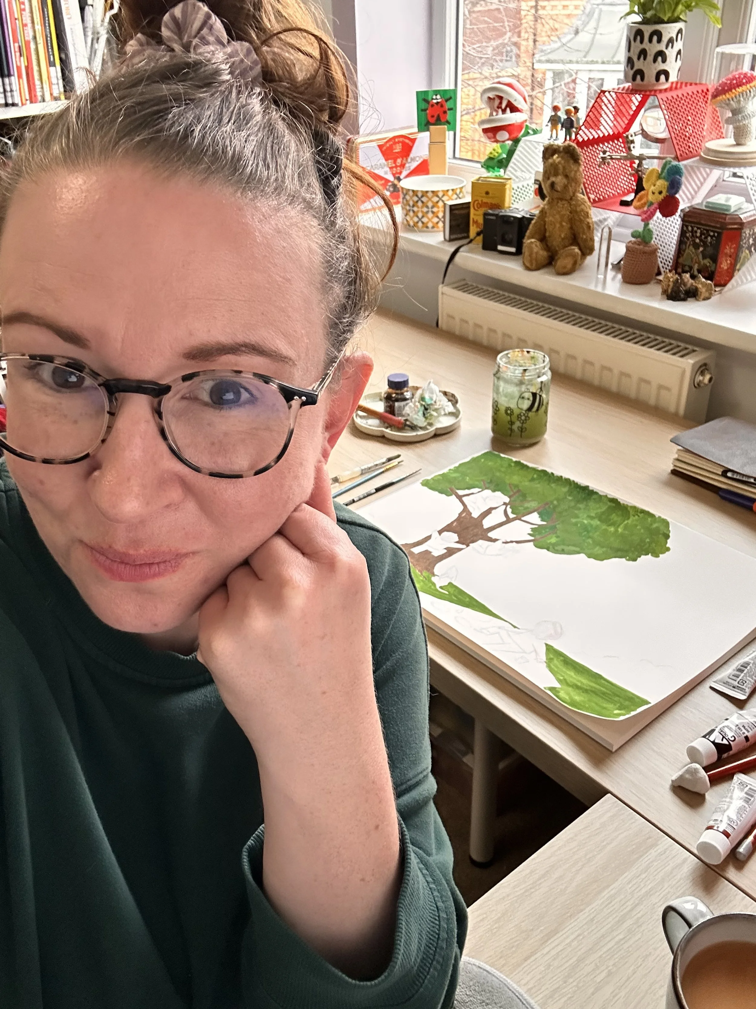A woman with glasses sitting at a desk next to an unfinished watercolor painting of a tree, with art supplies and a jar on the desk, and a windowsill with toys, books, and decorations in the background.