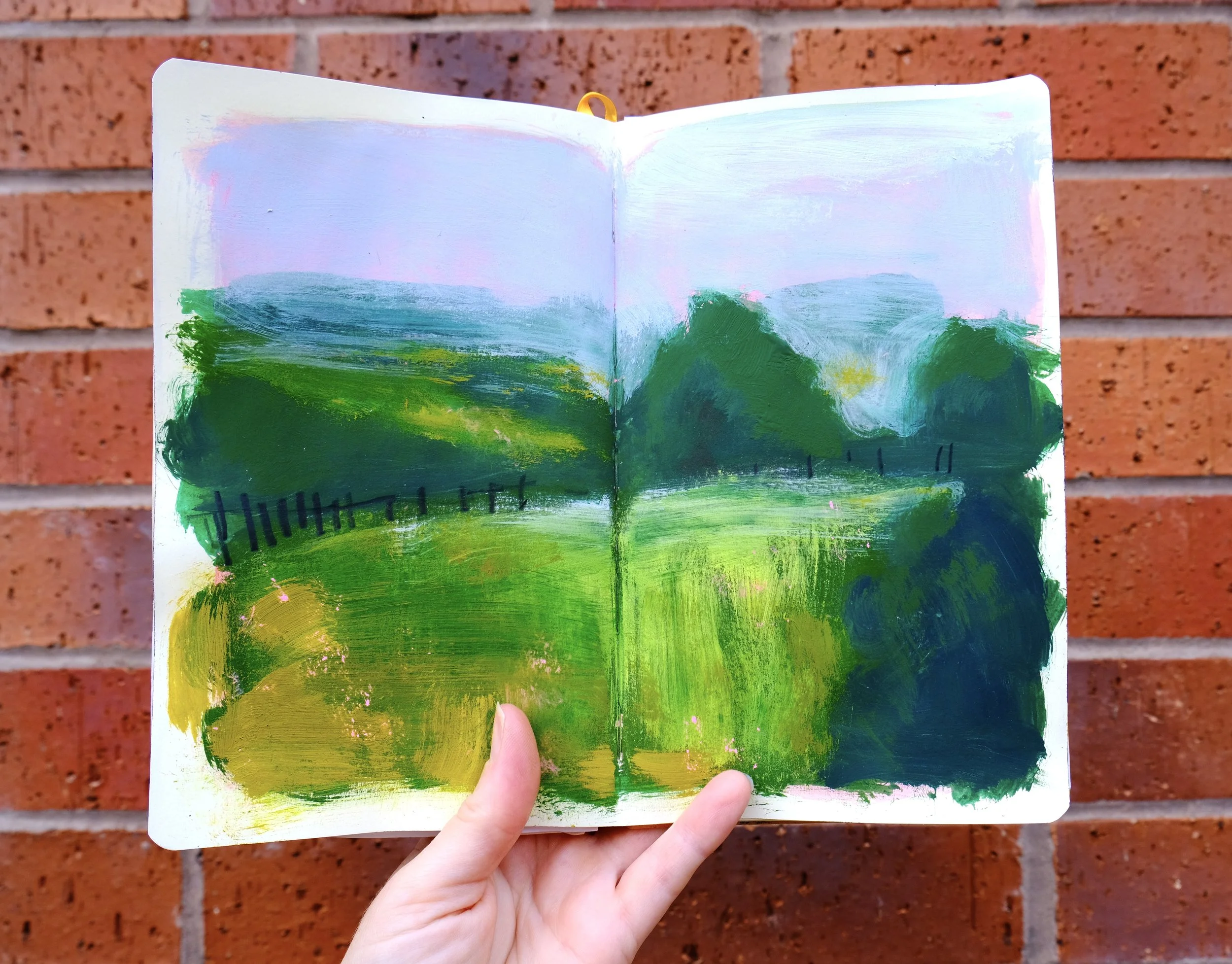 An open sketchbook with a colorful landscape painting of green fields, trees, and a blue sky, held against a brick wall.