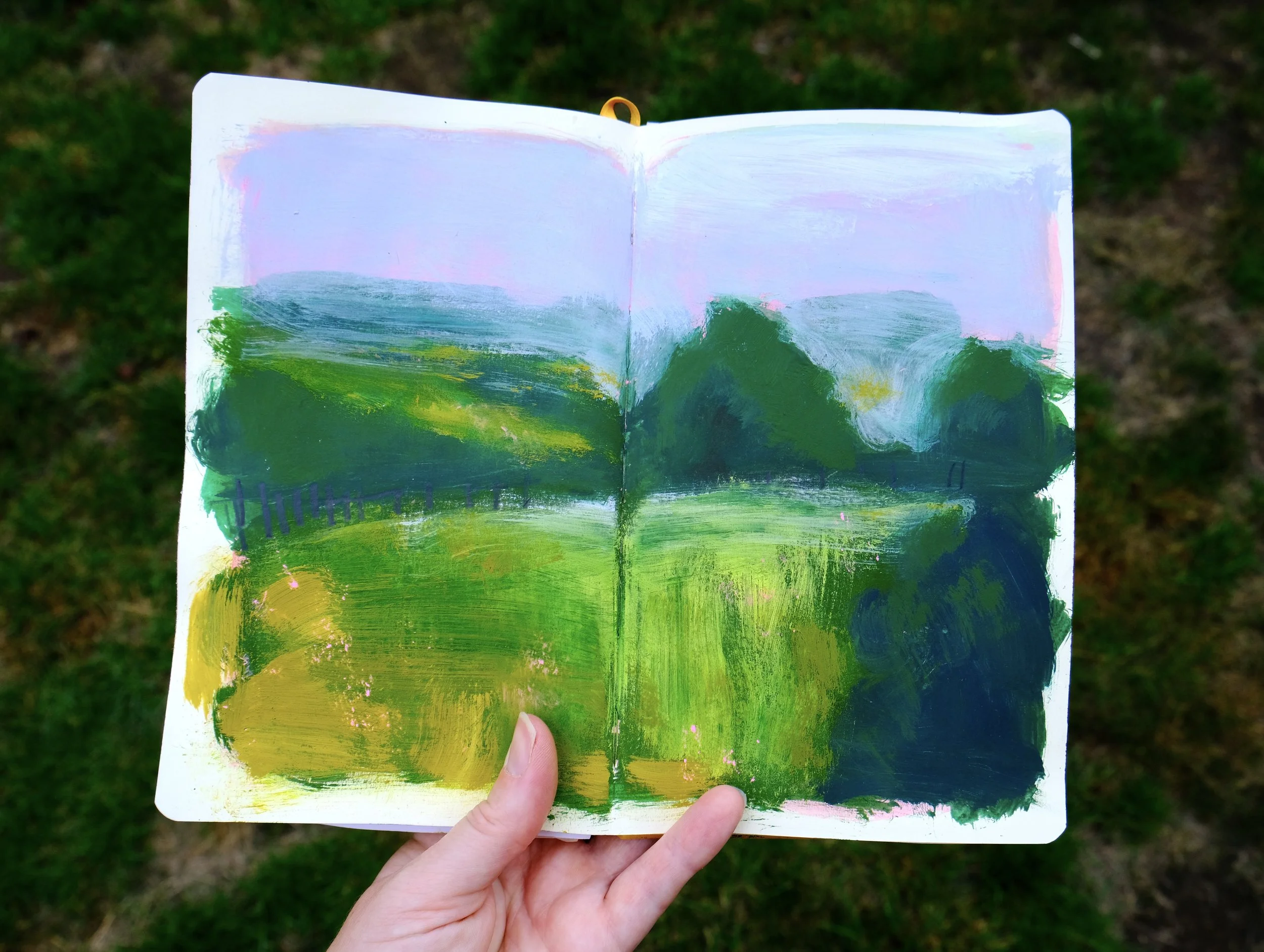 A hand holds an open sketchbook with an abstract landscape painting. The painting features green hills, blue trees, a pinkish sky, and various shades of foliage, created with broad, expressive brushstrokes.