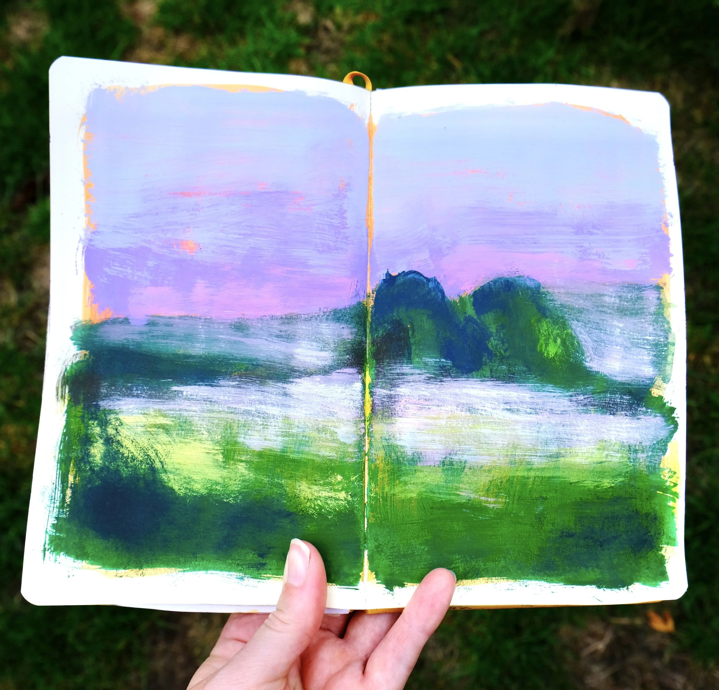 Hand holding an open sketchbook with a landscape painting of mountains, water, and trees during sunset.