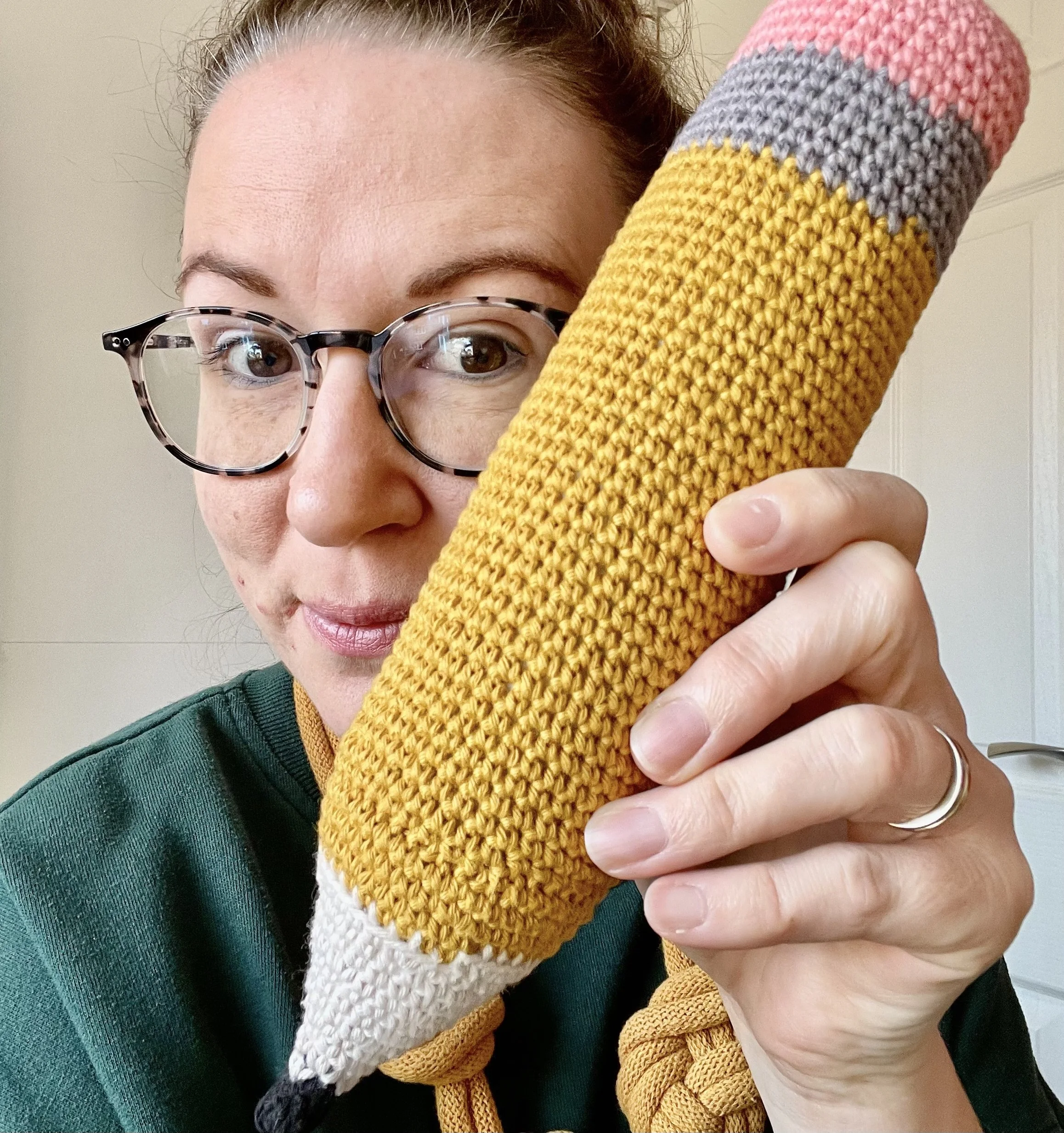 A woman with glasses holding a knitted colorful tomato-shaped object close to her face, indoors with a light-colored wall in the background.