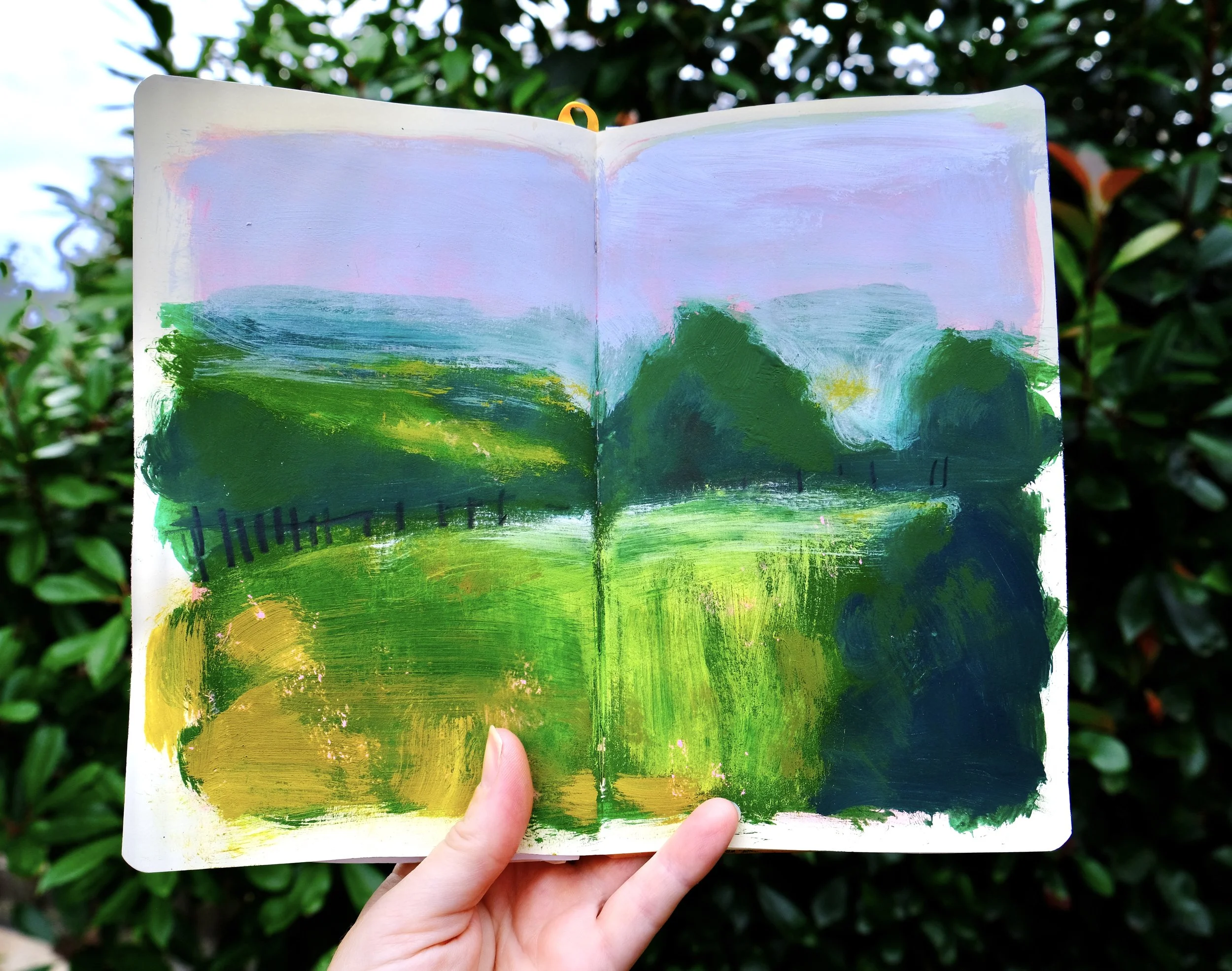 An open sketchbook with a painted landscape scene, held by a person's left hand against a background of green bushes.
