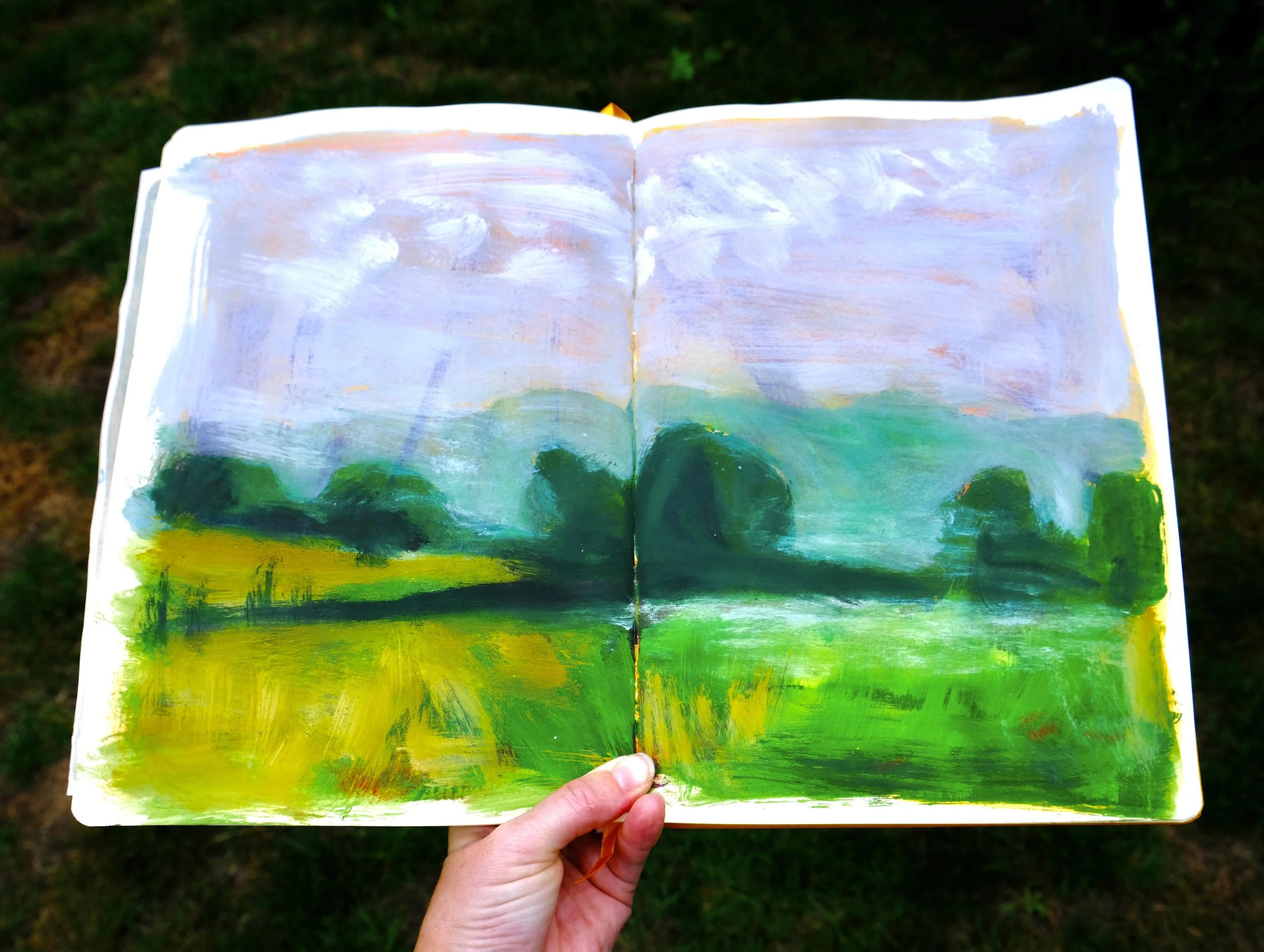 Hand holding a painted landscape sketchbook outdoors, depicting green fields, trees, and a cloudy sky.
