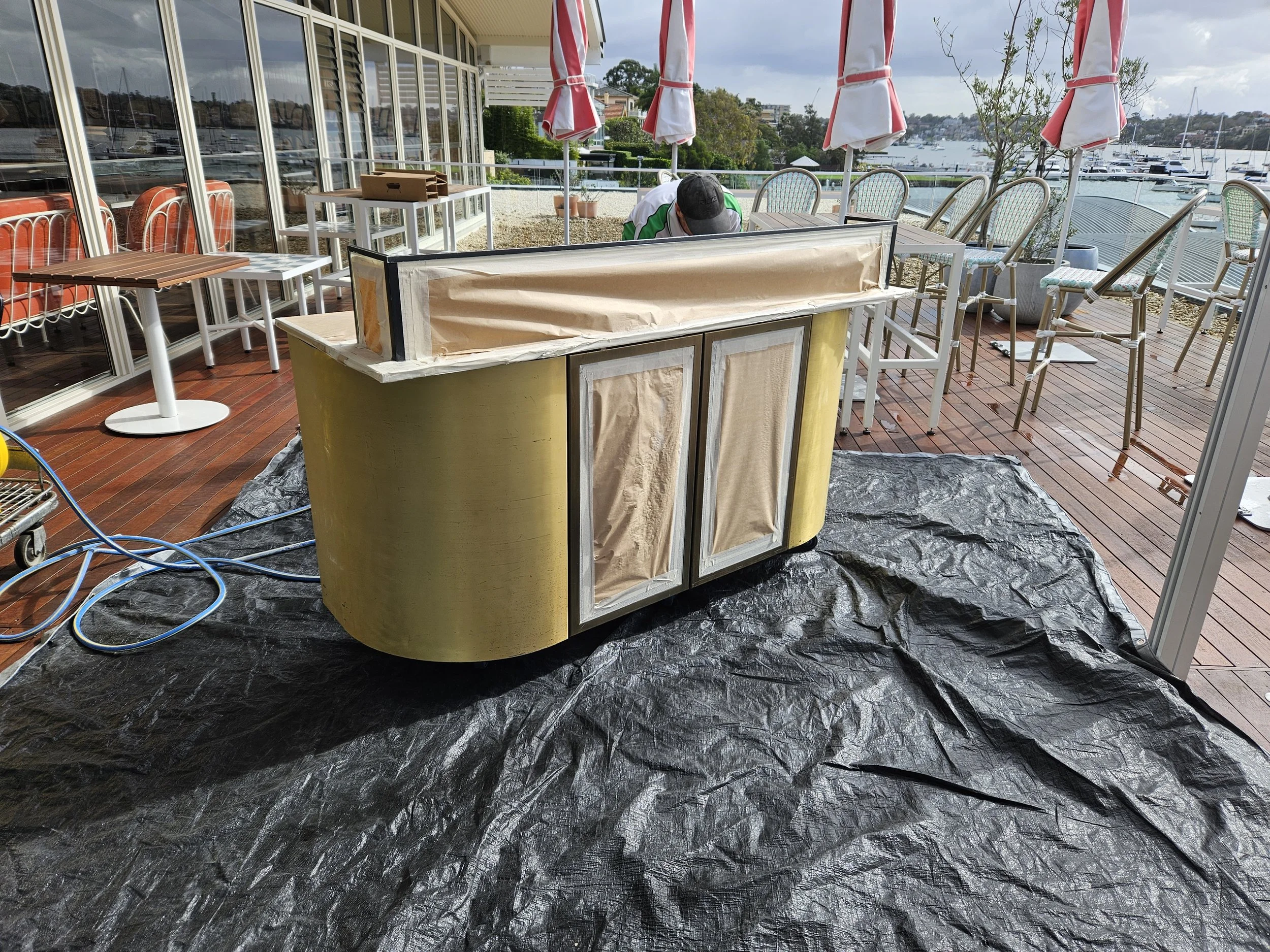 A partially covered green bar on a deck overlooking a marina with boats, with outdoor chairs and umbrellas, and a person working in the background.