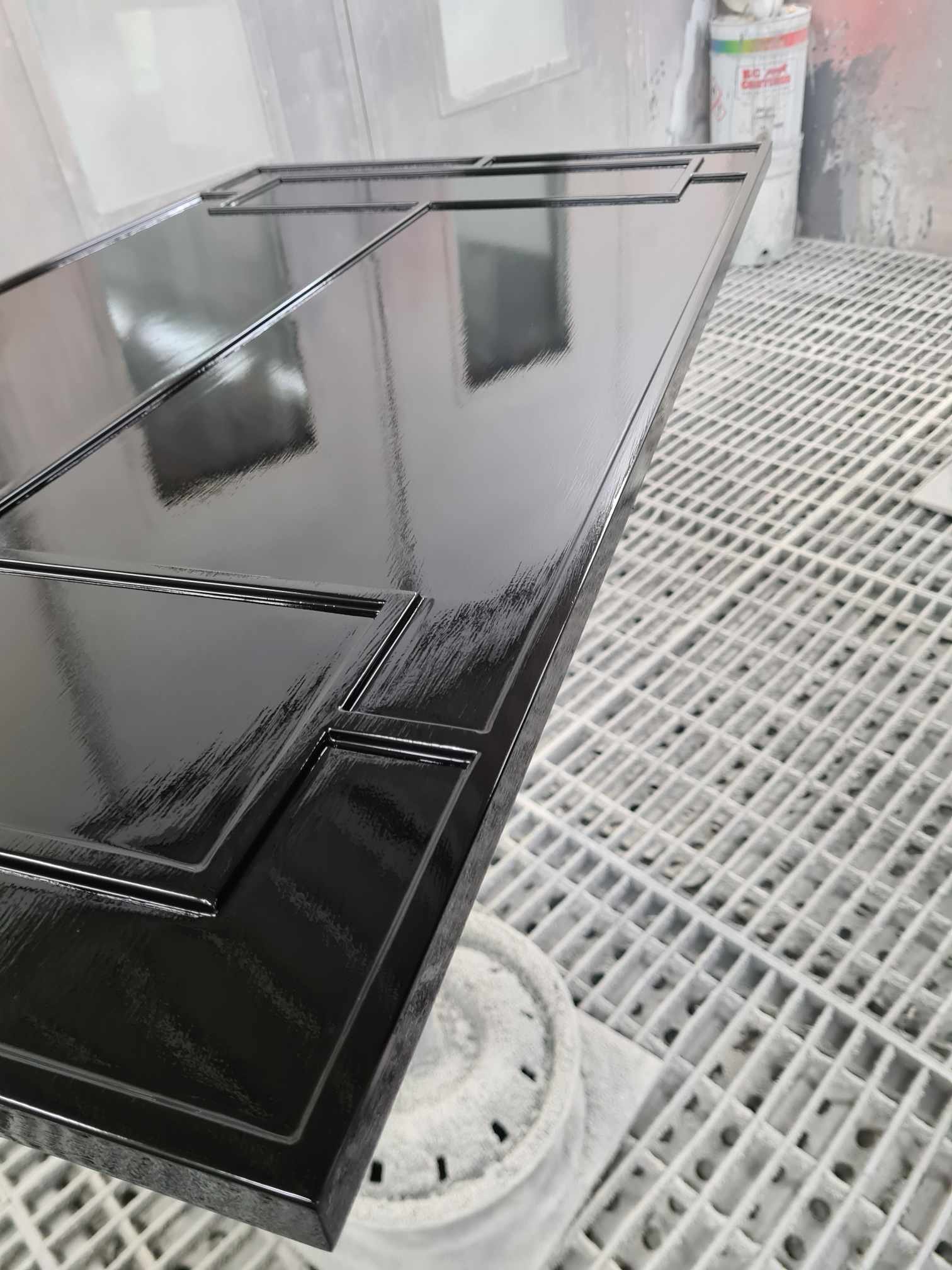 A black painted wooden cabinet door inside a spray booth, with a glossy finish, positioned on a white grate surface.