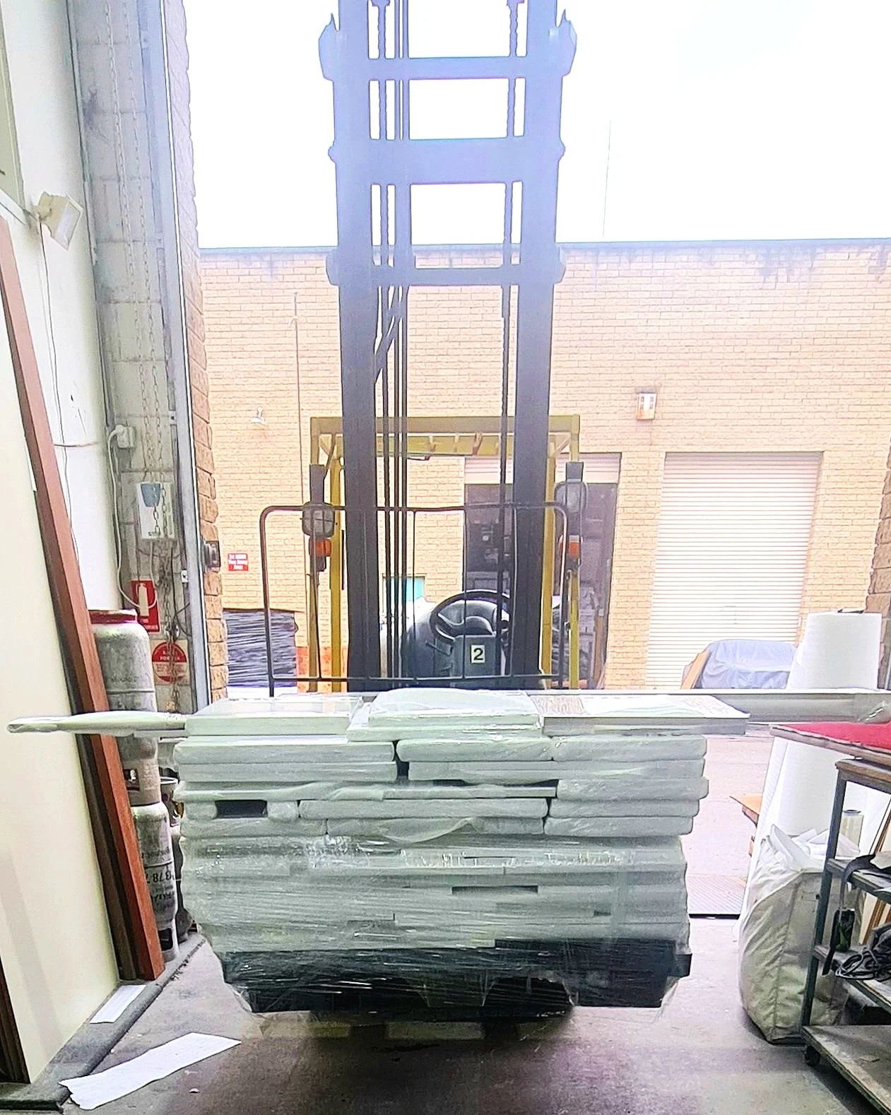 A forklift parked inside a warehouse near an open garage door, with stacked white foam boards on a pallet in front of it.